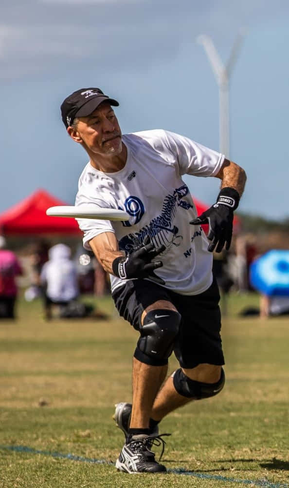 活力長者運動員在Android Ultimate Frisbee背景中 壁紙 — 一名中年男子穿著運動裝備，在陽光明媚的草地上向前撲去接住一個白色飛盤，背景有風力發電機。