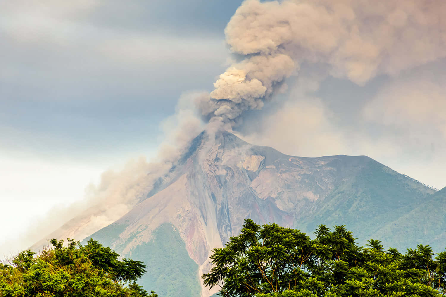 活火山上的日出 壁紙 — 火山爆發，濃煙滾滾升入多雲的天空，籠罩在茂密的綠色樹梢之上。