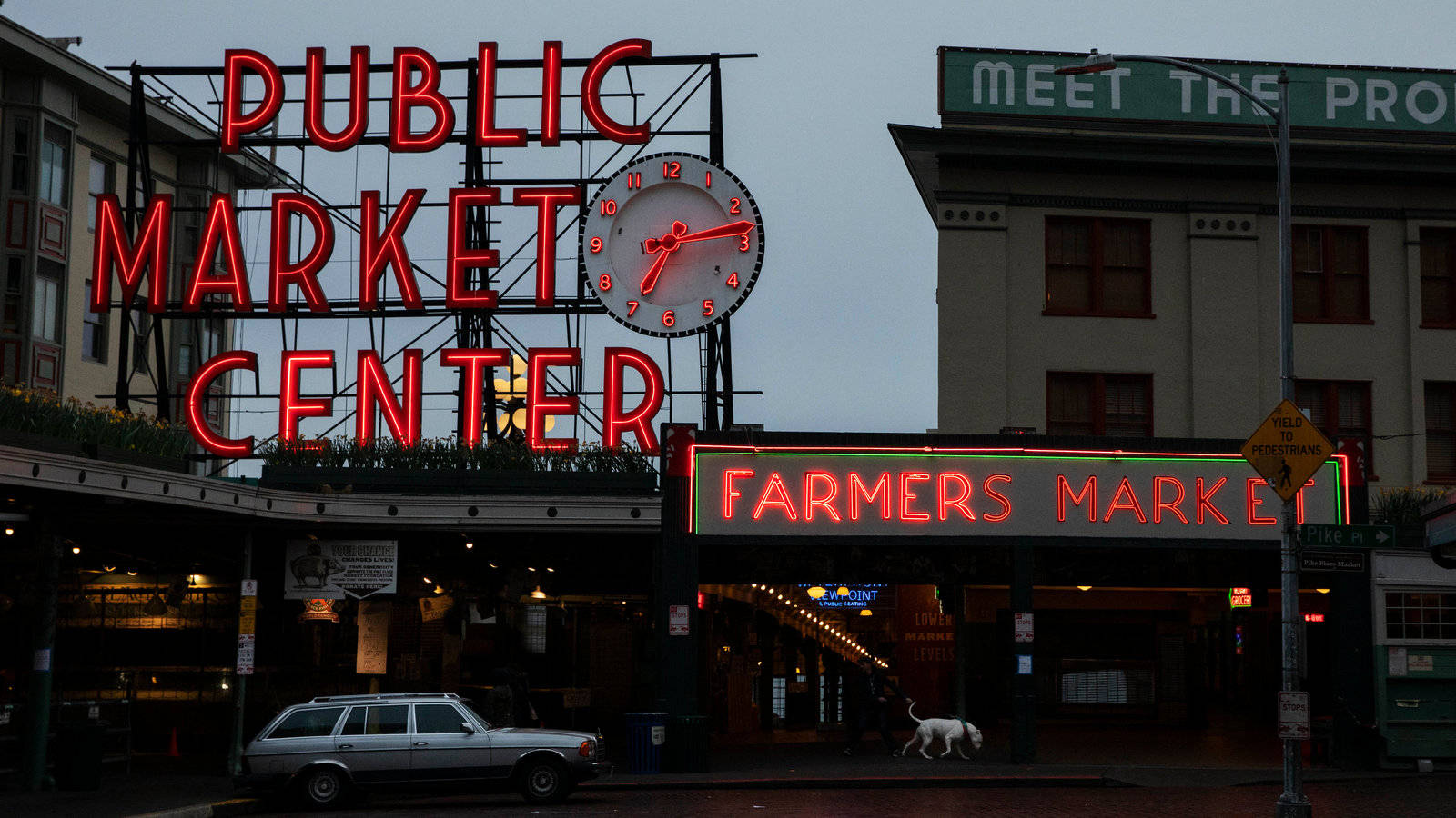 Public Market Center和Farmers Market的霓虹燈招牌在黃昏時發光，附近有一輛銀色汽車和一隻狗。