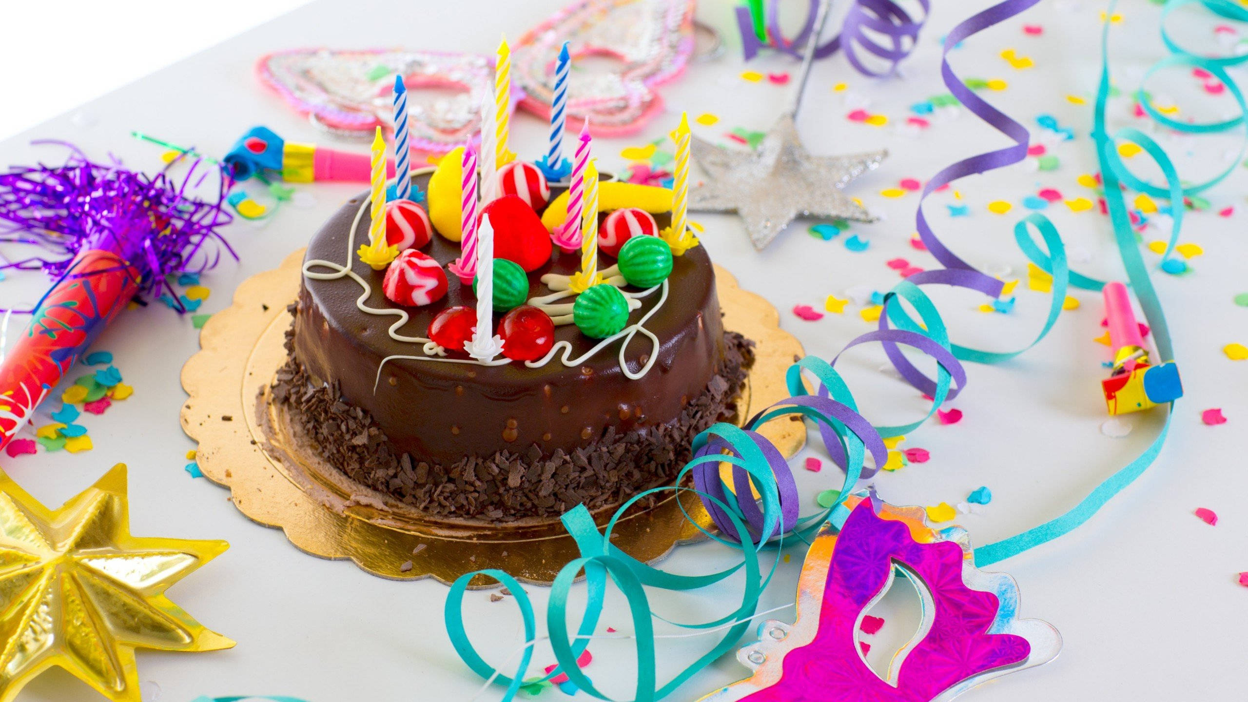 派對用品裝飾生日蛋糕 壁紙 — 一個朱古力生日蛋糕，上面有彩色糖果和蠟燭，周圍是派對五彩紙屑彩帶和裝飾。