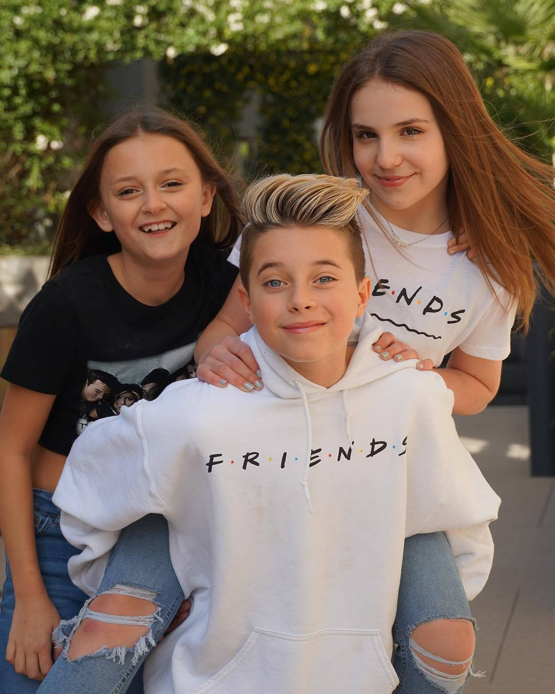 三位微笑的青少年穿著 Friends 連帽衫和破洞牛仔褲在戶外擺姿勢。