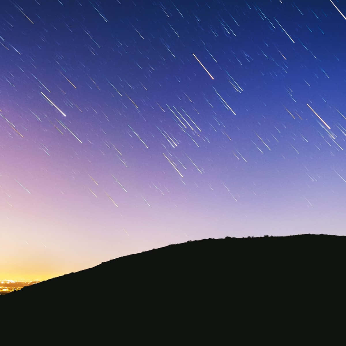 流星劃破夜空的壯麗景象 壁紙 — 星軌劃過暮色天空，在黑暗山丘剪影之上，遠方城市燈光在地平線閃爍。