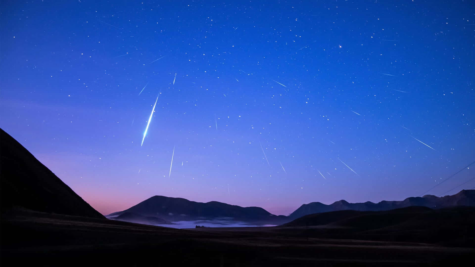流星劃過繁星滿天的夜空 壁紙 — 一顆明亮的流星劃過佈滿星空的暮色天空，下方是輪廓分明的山脈和霧氣瀰漫的山谷。