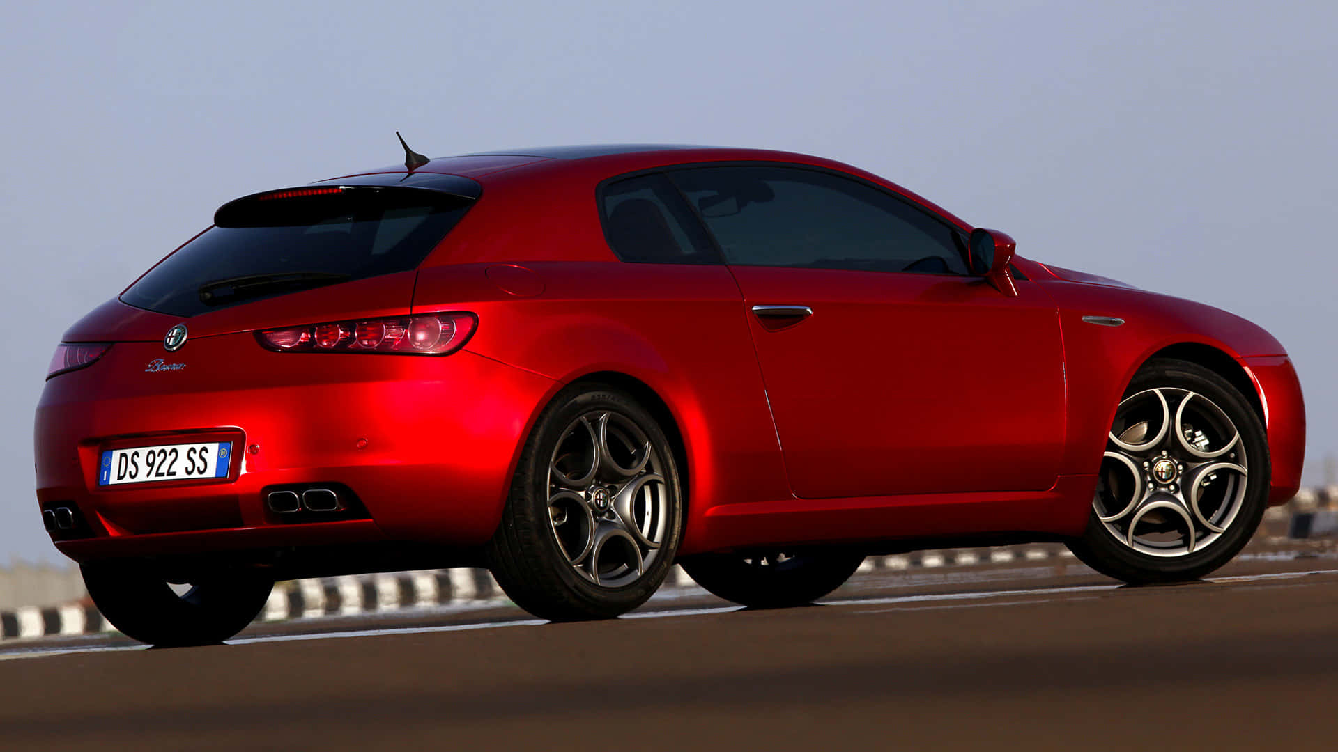流線型 Alfa Romeo Brera 於壯麗戶外景觀中 壁紙 — 一輛光澤的紅色愛快羅密歐Brera運動轎跑車，沿著陽光明媚的高速公路行駛，擁有流暢的曲線和合金輪圈。