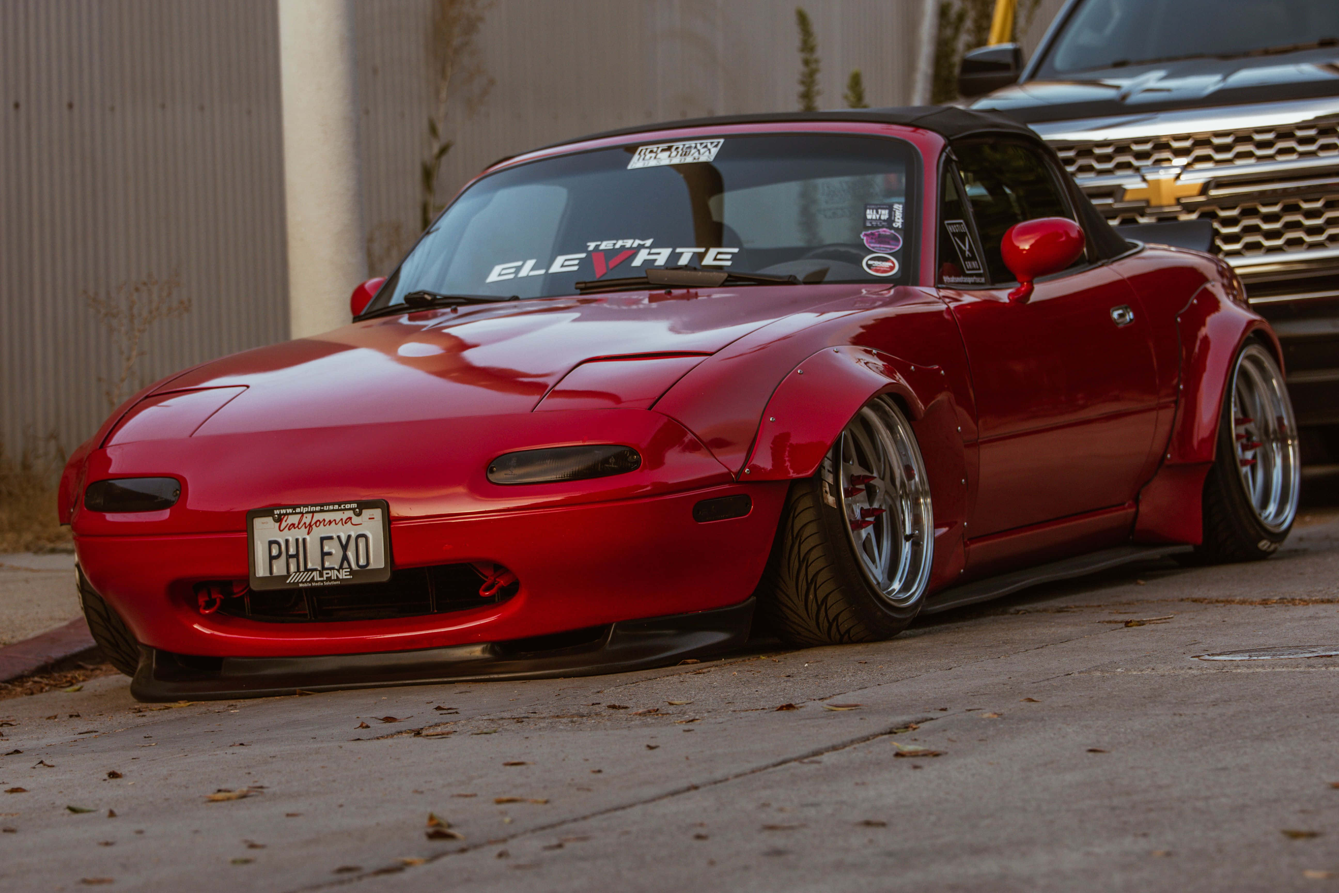 流線型1995年紅色 Mazda MX-5 Miata 壁紙 — 一輛光澤紅色改裝萬事得MX-5，配備寬體套件和訂製輪圈，停泊在建築物附近的混凝土停車場。