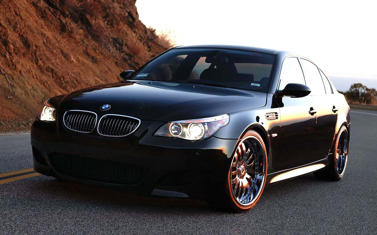 流線型BMW M5疾馳於風景路線 壁紙 — 一輛亮黑色BMW M5轎車配備定制輪轂，在黃金時段沿著蜿蜒的道路閃耀，背景是崎嶇的懸崖。