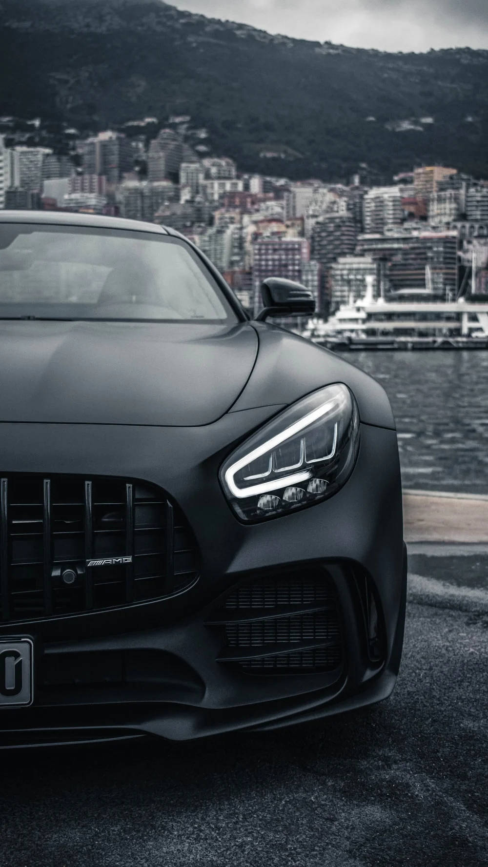 流線型黑色AMG GT 壁紙 — 一架啞黑色平治AMG跑車停泊在海濱，背景有城市景觀和山脈。