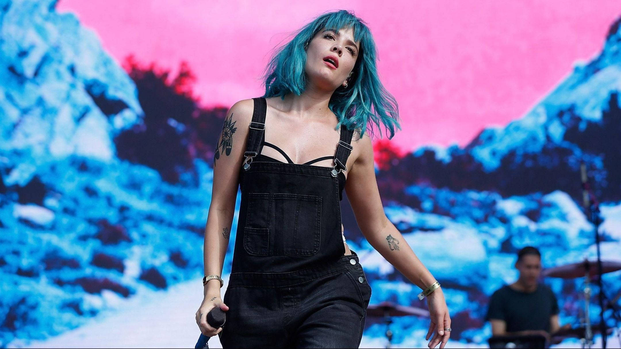 流行天后Halsey於Billboard音樂節閃耀登場 壁紙 — 一位擁有充滿活力的藍色頭髮的表演者在舞台上唱歌，背景是鮮豔的粉紅色和藍色山脈。