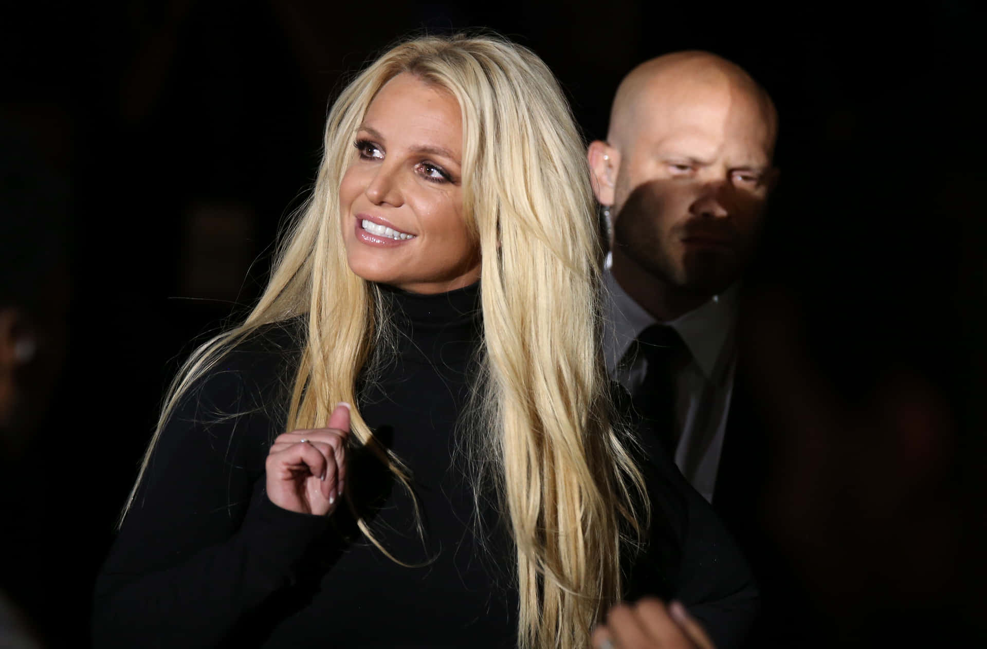 Britney Spears 穿著黑色高領毛衣微笑，身後有穿西裝的男子。