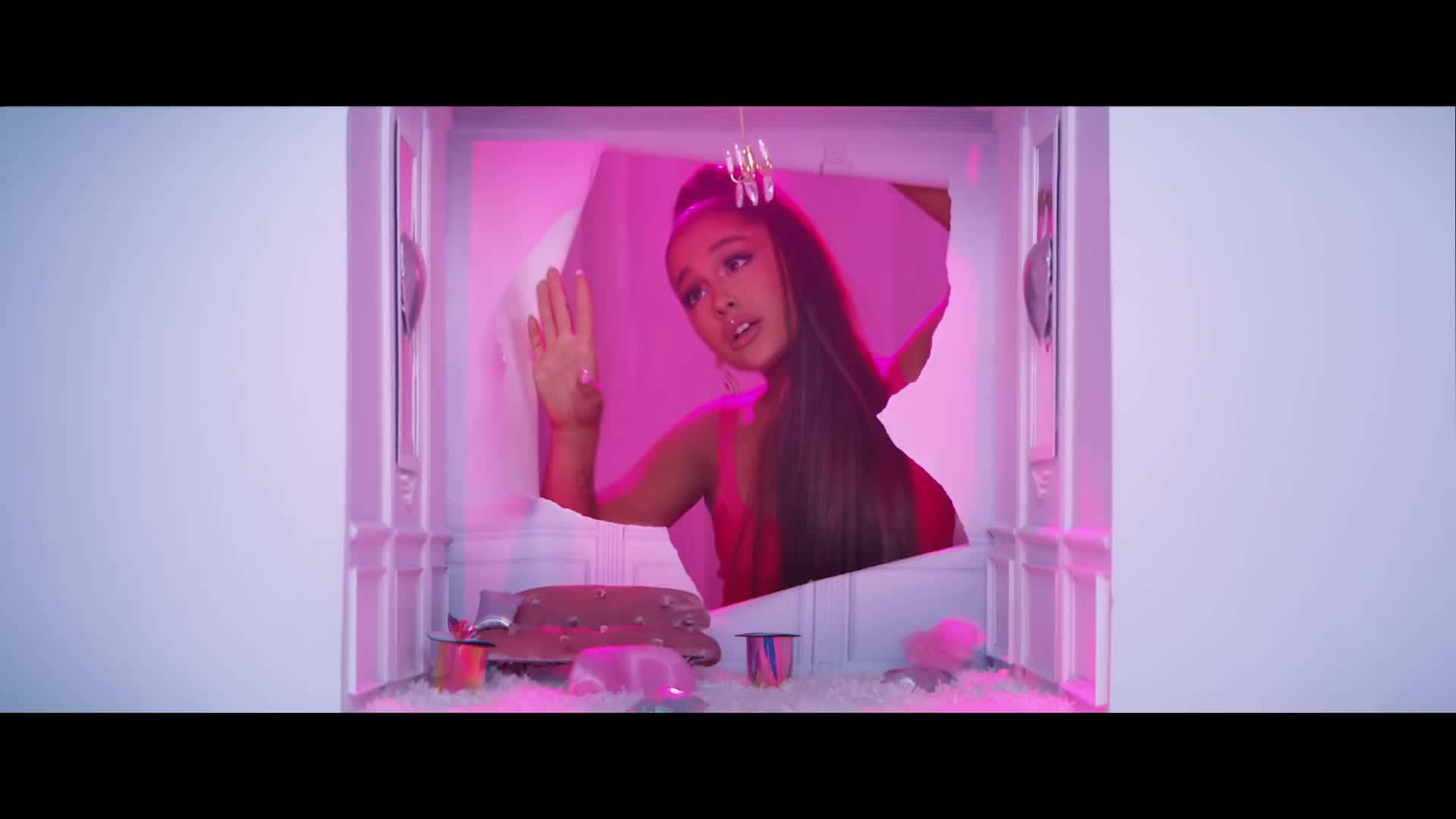 Ariana Grande 喺一個鋪滿白色絨毛地板、粉紅色燈光嘅娃娃屋房間入面。