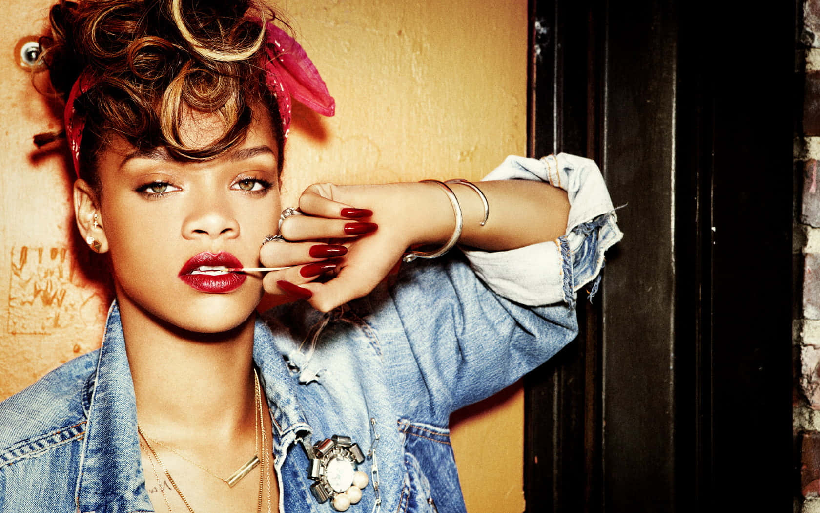 Rihanna 身穿牛仔外套，塗上紅色唇膏和戴頭帶，靠牆擺姿勢。