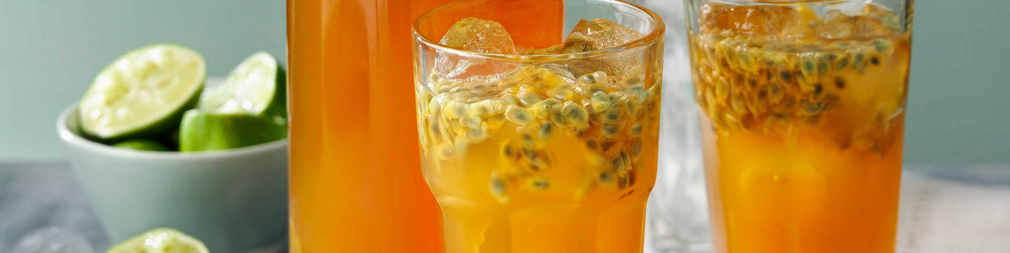 浩瀚海洋上的迷人日出 壁紙 — 兩杯裝滿鮮豔橙色熱情果飲料和冰塊的玻璃杯，旁邊放著一碗切片青檸，置於灰色表面上。