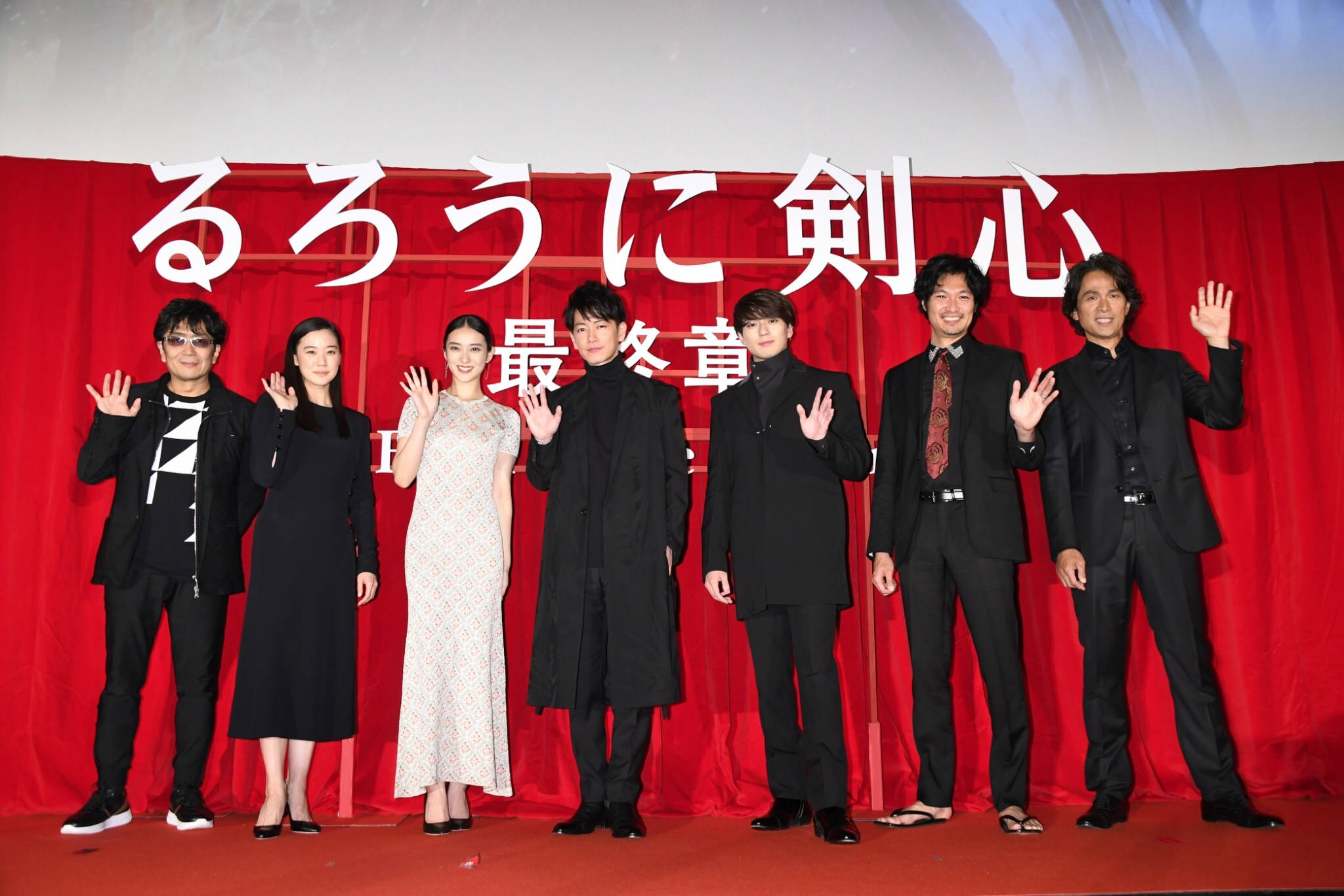 浪客劍心 最終篇 演員陣容 壁紙 — 七位演員在紅地毯上向鏡頭揮手，背景是帶有白色日文字的紅色窗簾。