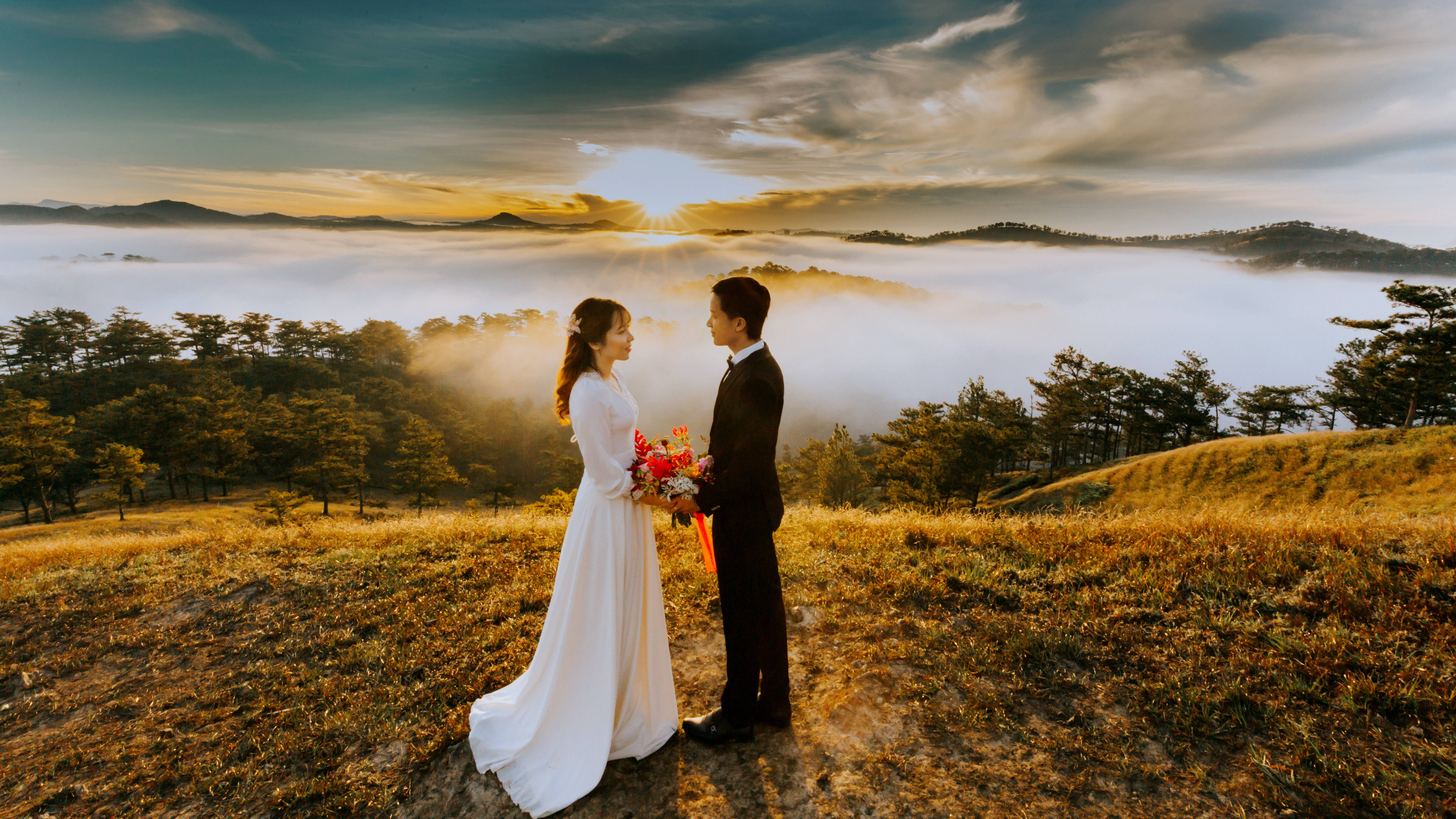 浪漫情侶山頂婚紗照 壁紙 — 新娘和新郎站在日出時陽光照耀的山頂上，凝視著彼此的眼睛，薄霧在下方翻滾。