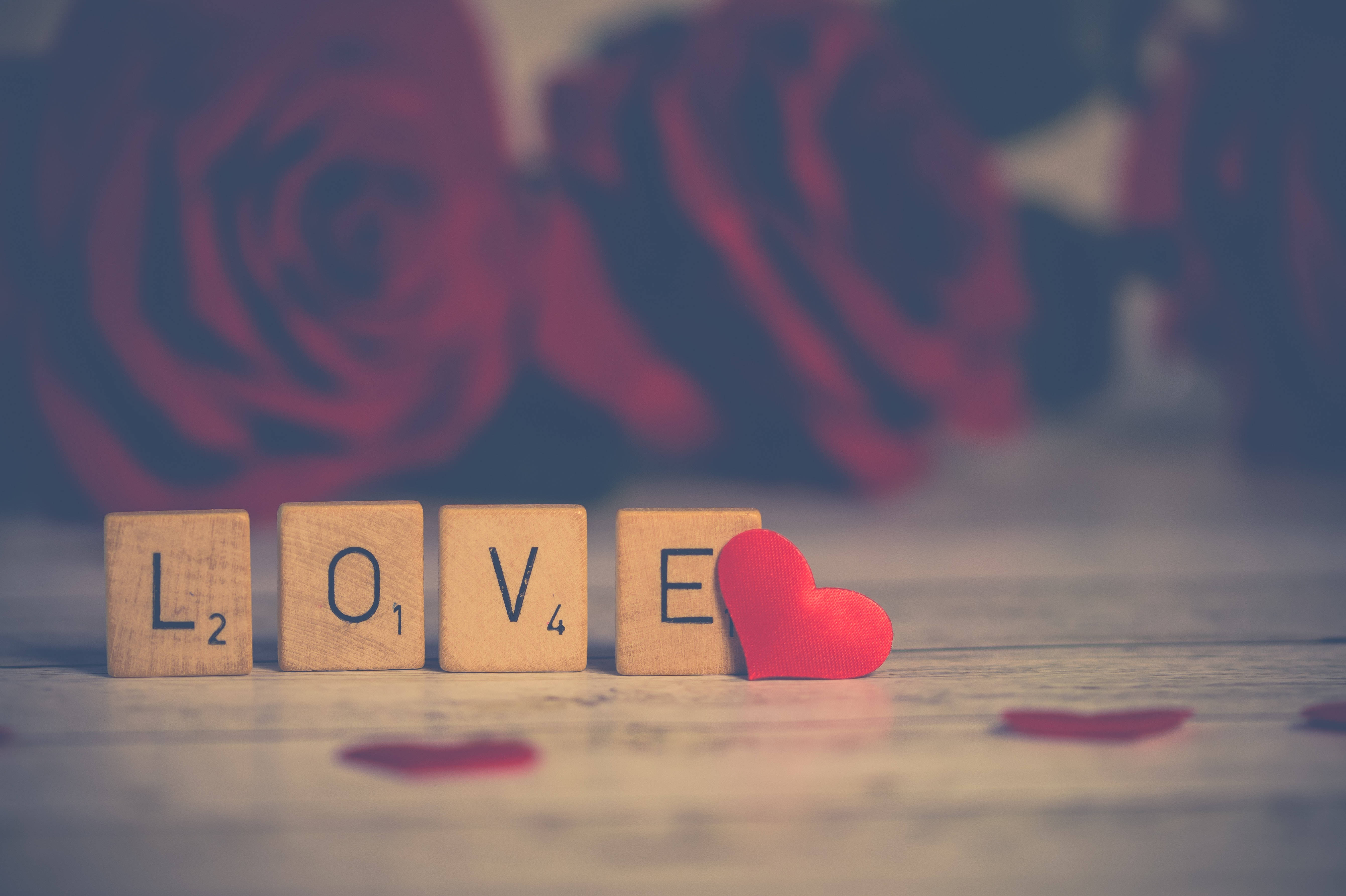 浪漫愛意拼字遊戲 壁紙 — 木製 Scrabble 字母拼出 LOVE，旁邊有一個紅心，背景是模糊的紅玫瑰。