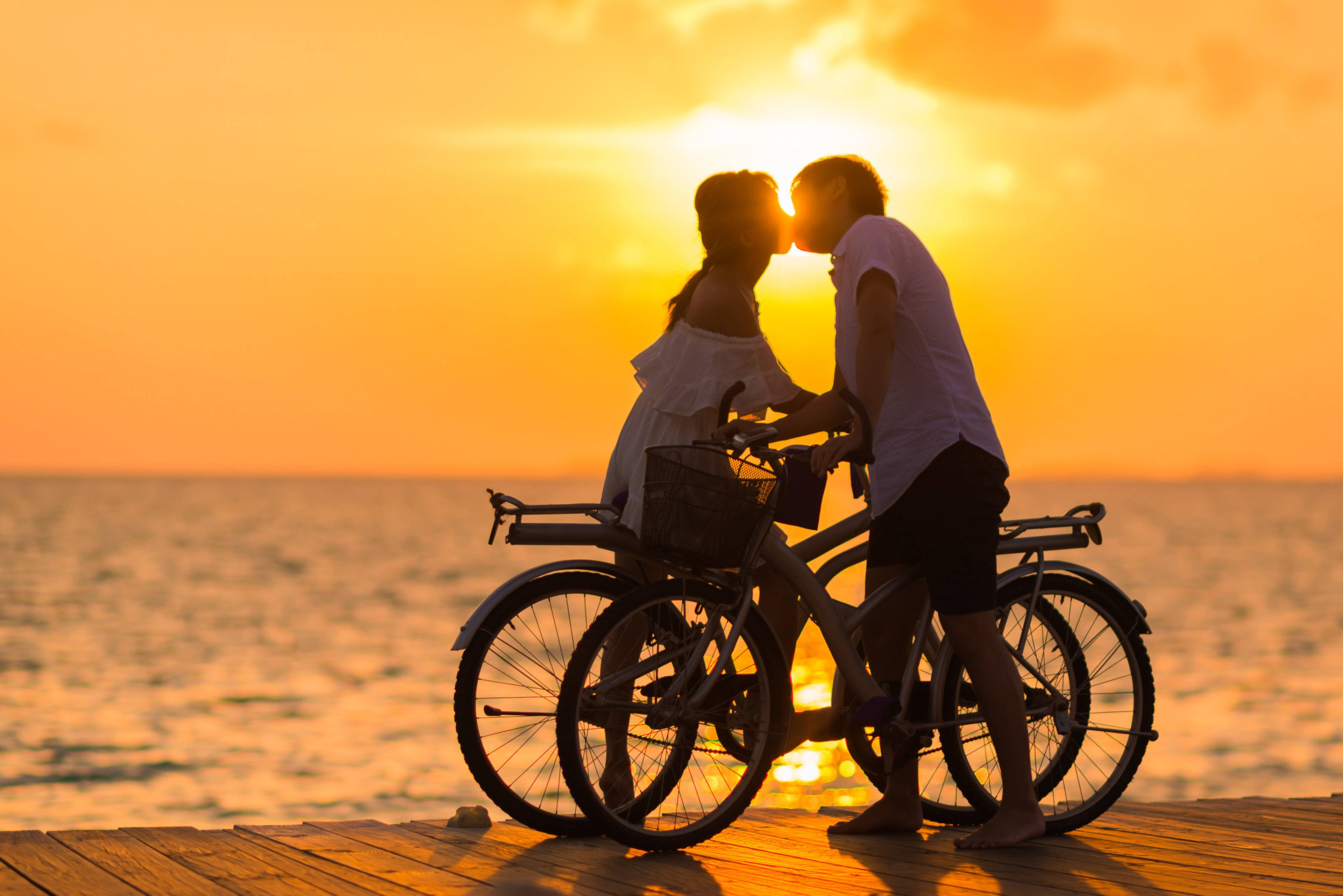 浪漫愛戀：夕陽下的自行車情侶 壁紙 — 一對情侶在日落時分的木製碼頭上接吻，單車放在旁邊，剪影映襯著金色的天空和海洋。