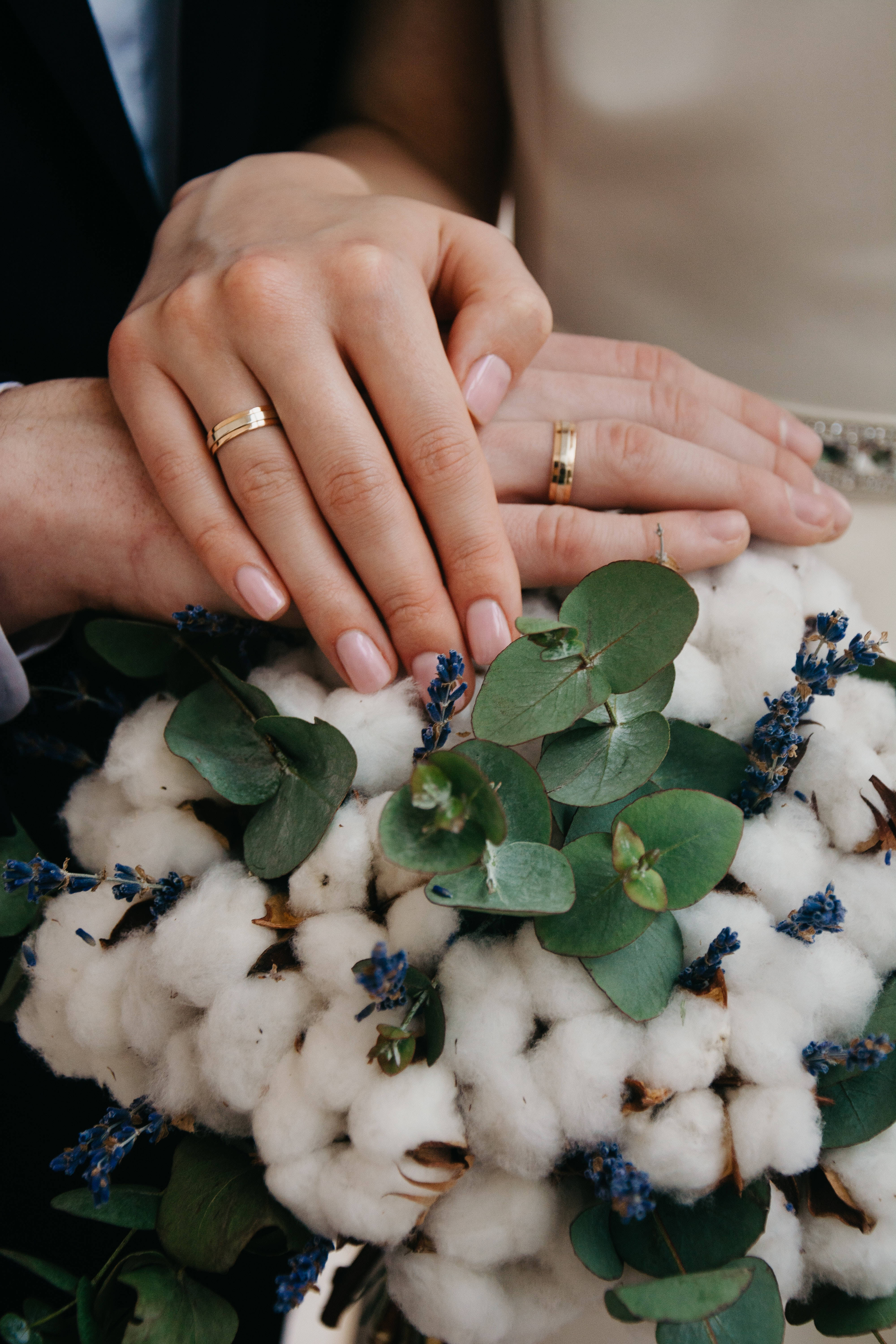 浪漫愛戀：婚戒交握 壁紙 — 兩隻戴著結婚戒指的手輕輕地放在一個柔軟的棉花花束上，花束點綴著綠色桉樹和小藍花。