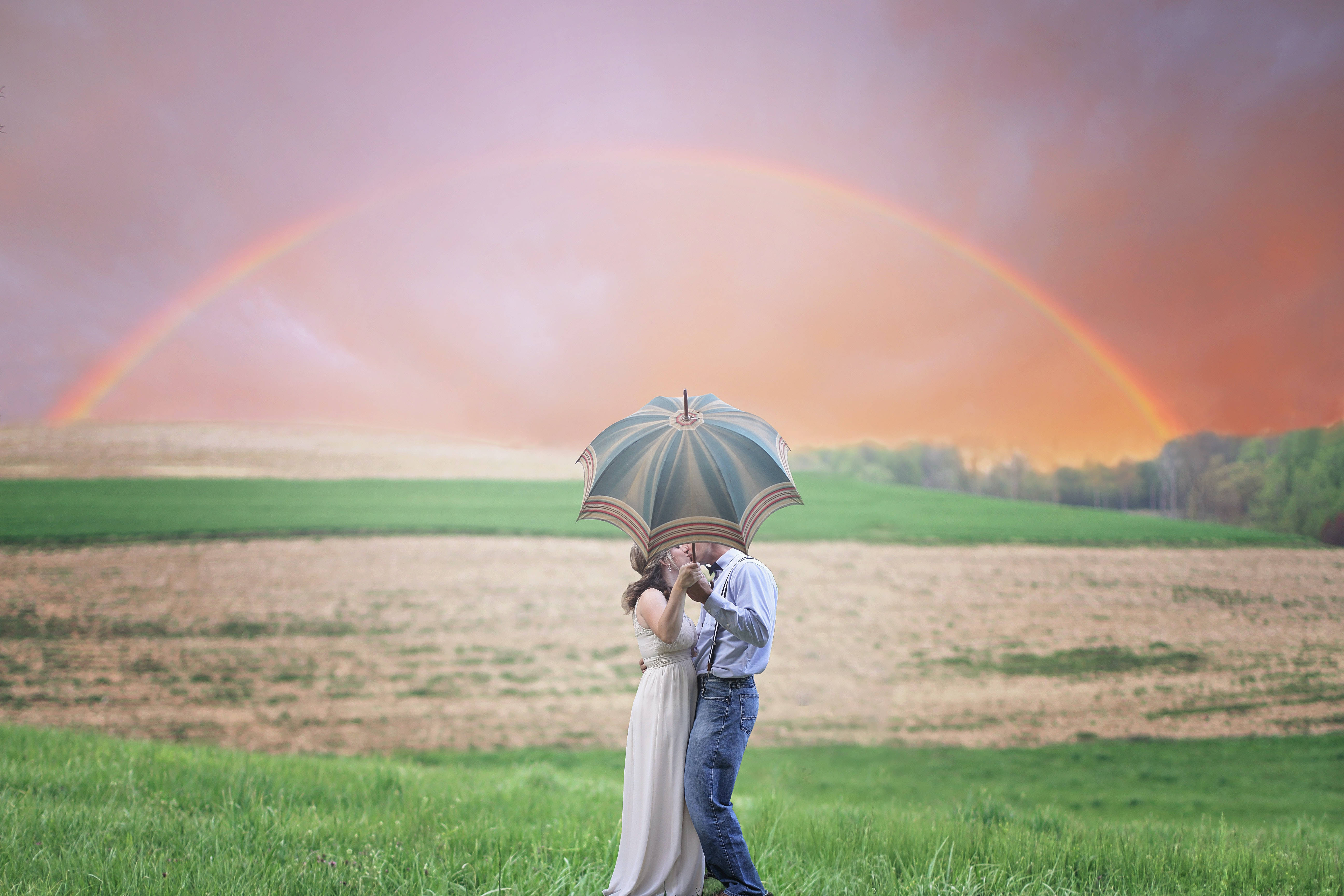 浪漫愛戀：彩虹下的情侶 壁紙 — 一對情侶在田野中，於圖案雨傘下接吻，一道鮮豔的彩虹橫跨夕陽天空。