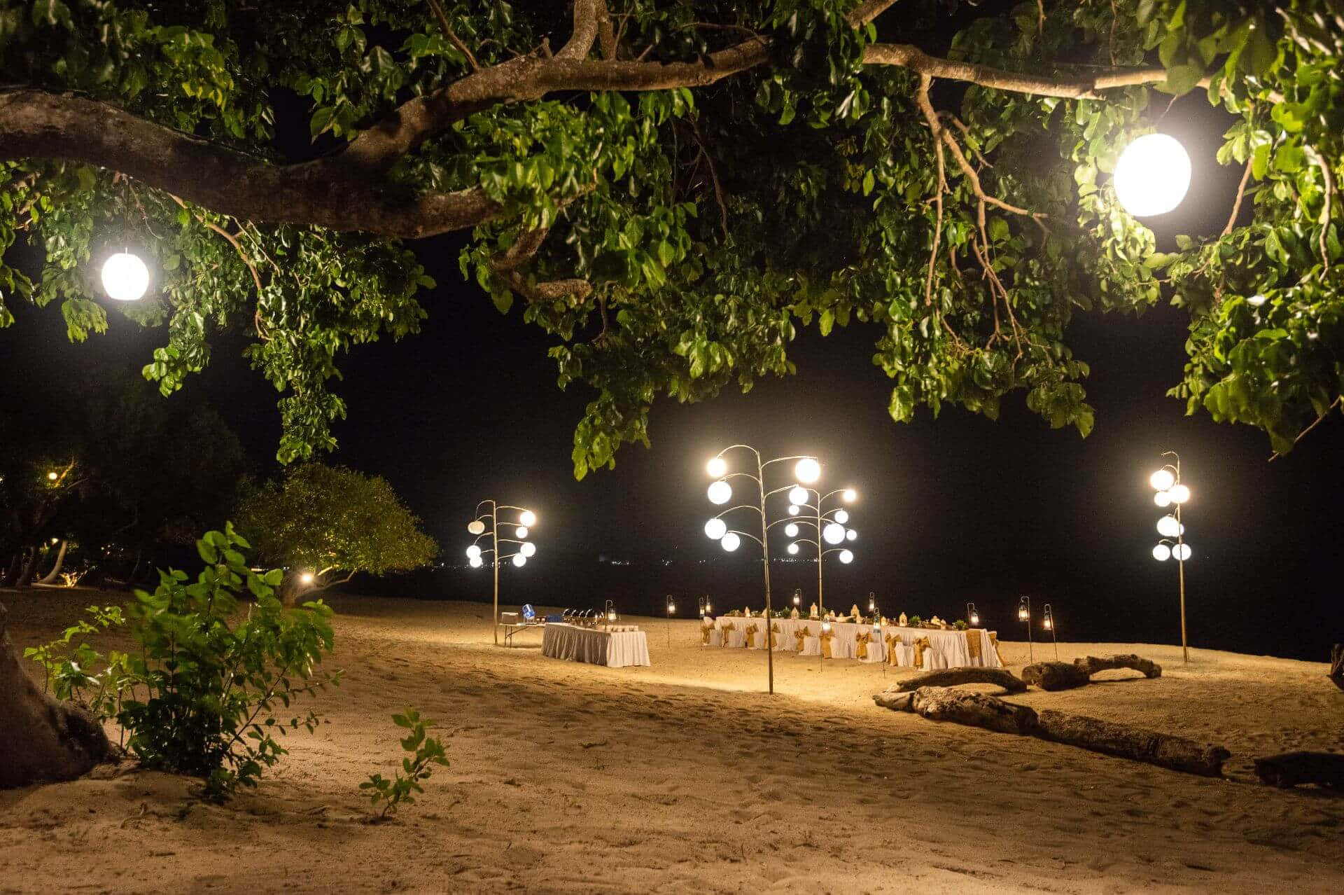 浪漫晚餐夜灘圖 壁紙 — 夜晚海灘婚禮佈置，在發光燈籠和枝葉茂盛的樹木下，在沙灘上投下柔和陰影。