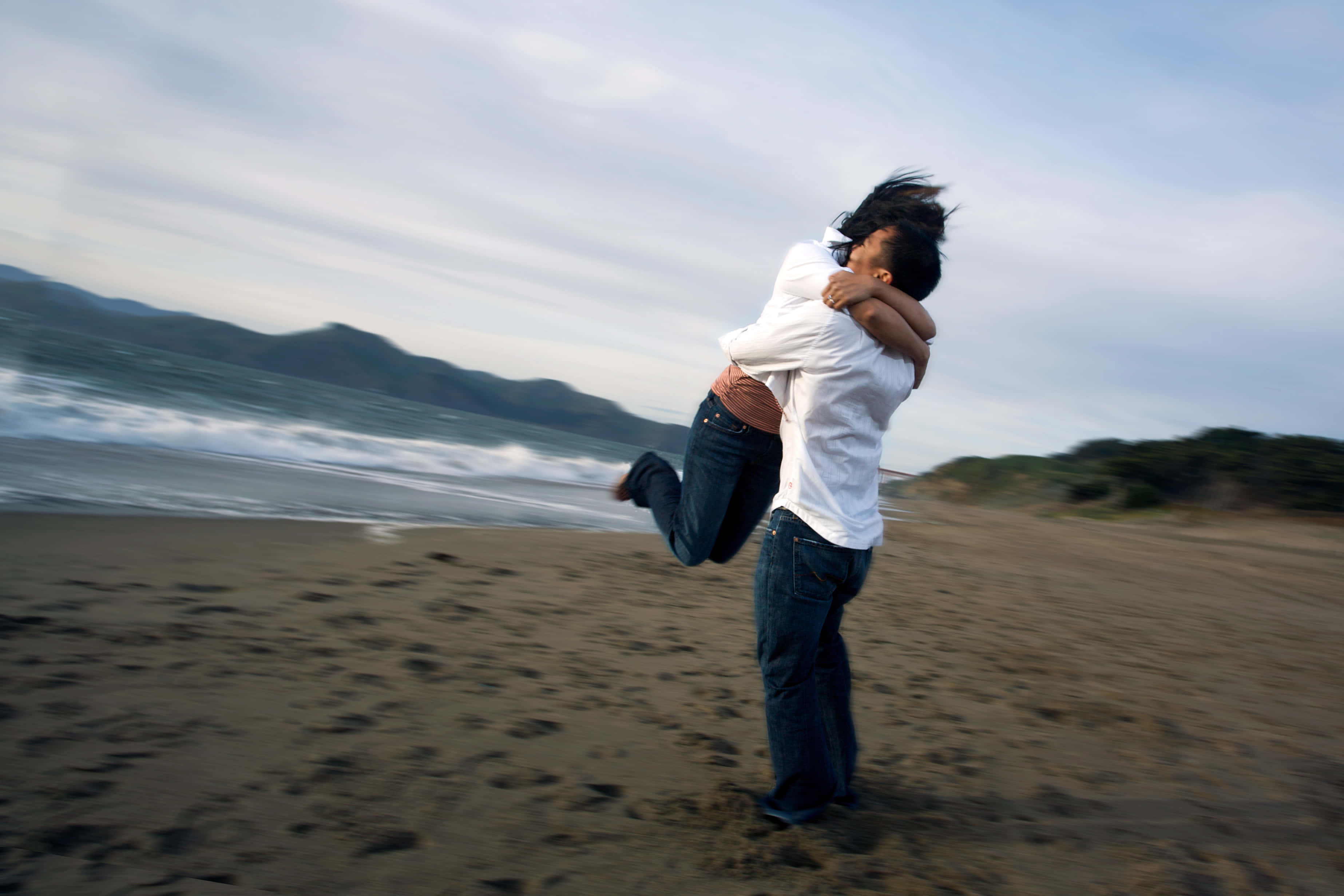 浪漫海灘擁抱畫面 壁紙 — 一個男人在風吹沙灘上，快樂地以動態擁抱舉起一個女人，海浪拍打，遠山可見。