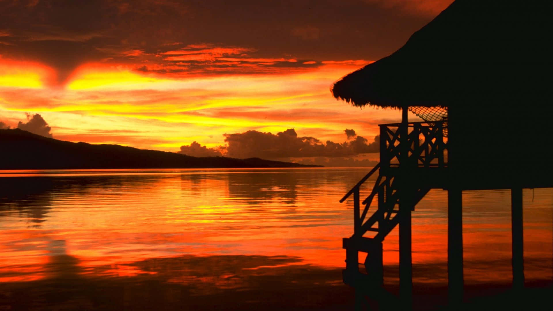 浪漫的海灘日落 壁紙 — 充滿活力的夕陽將天空和水面染成火熱的橙色和紅色，一座茅草屋的剪影矗立在水面上的高蹺上。