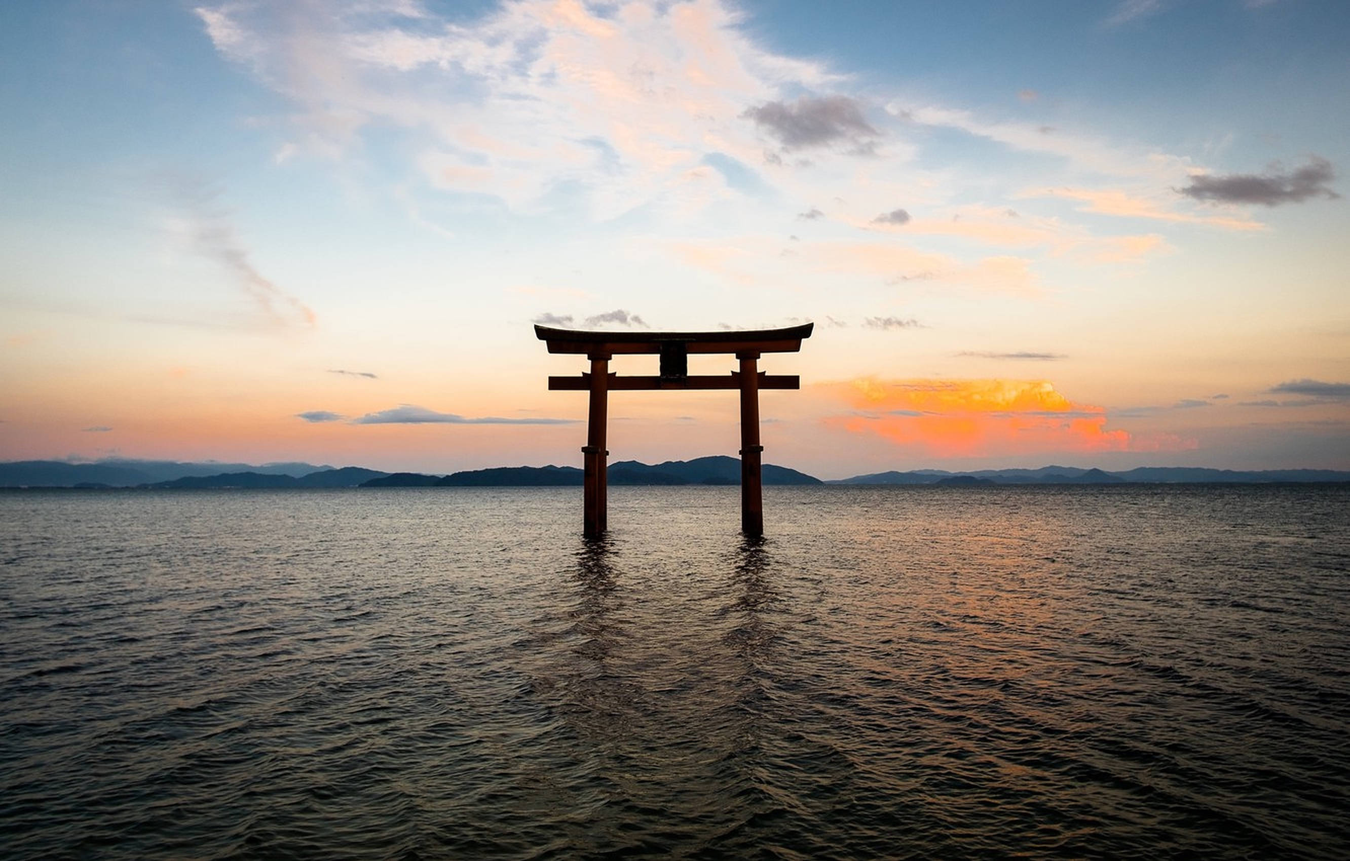 海中的鳥居門 壁紙 — 一座寧靜的木製鳥居矗立在平靜的水面上，夕陽西下，橙色的雲彩和遠山映襯在柔和的藍天下。