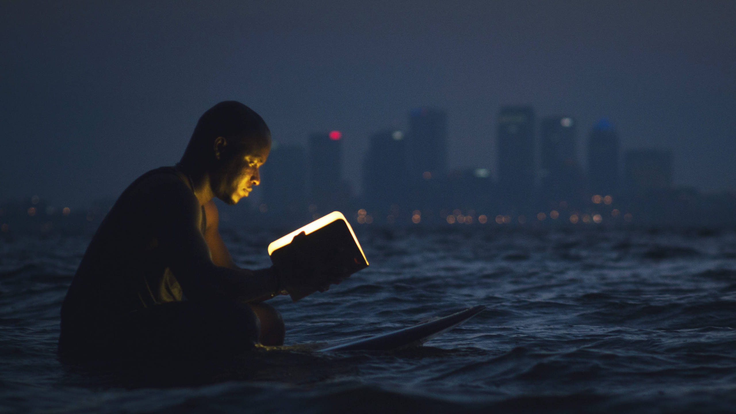 海中閱讀書籍的男子 壁紙 — 一個人夜晚坐在黑暗的水中，閱讀一本發光的書，背景是模糊的城市天際線。