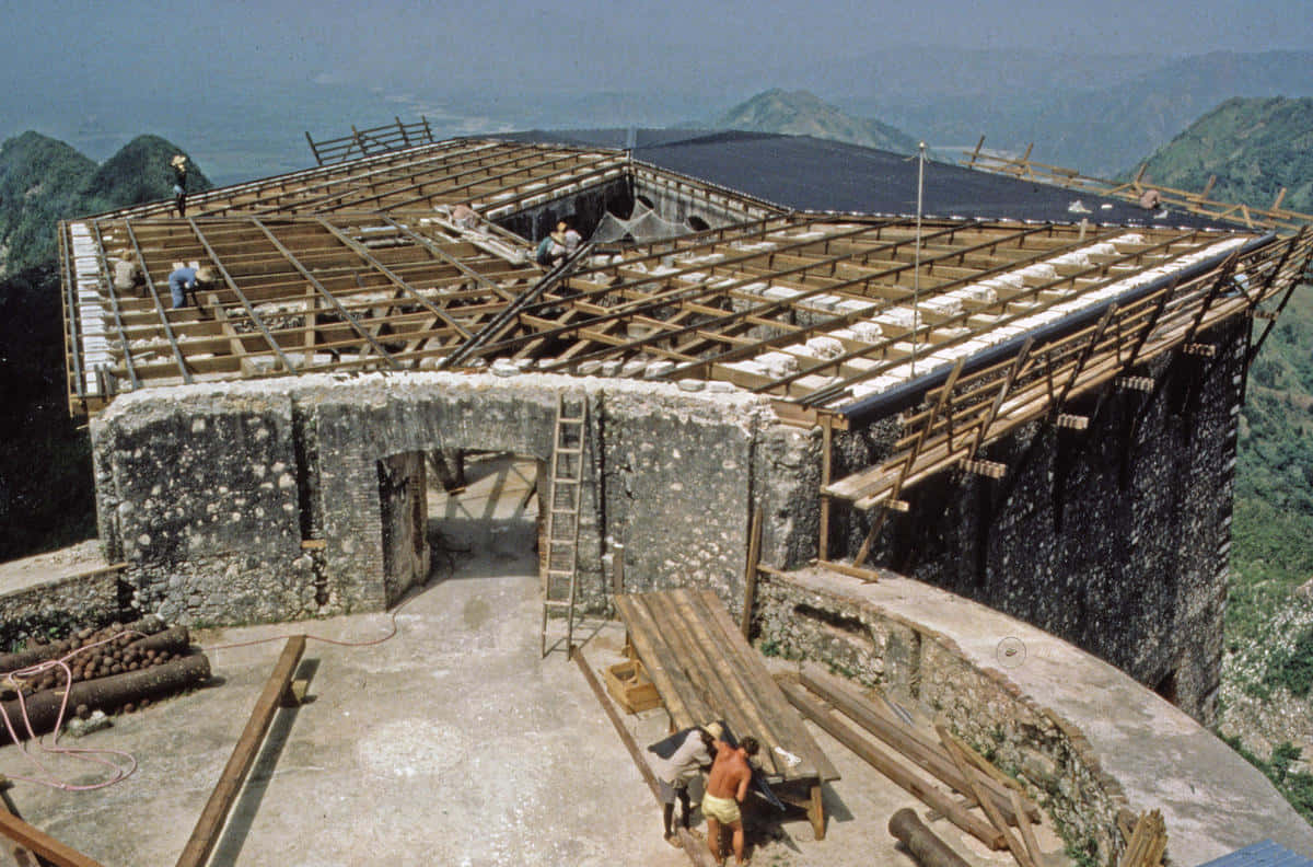 海地拉弗里耶爾城堡建設中 壁紙 — 工人在山頂的石建築上建造木屋頂，遠方有翠綠的山丘，天空朦朧。