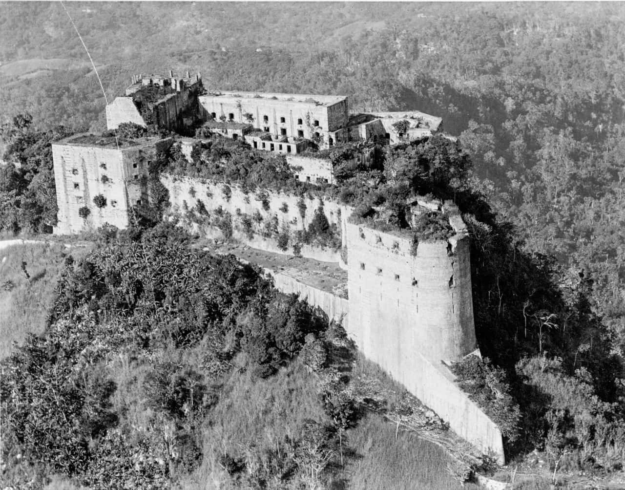 海地的壯麗拉弗里耶堡壘 壁紙 — 俯瞰一座風化的石頭堡壘，坐落在樹木繁茂的山丘上，城牆和塔樓長滿了植物。