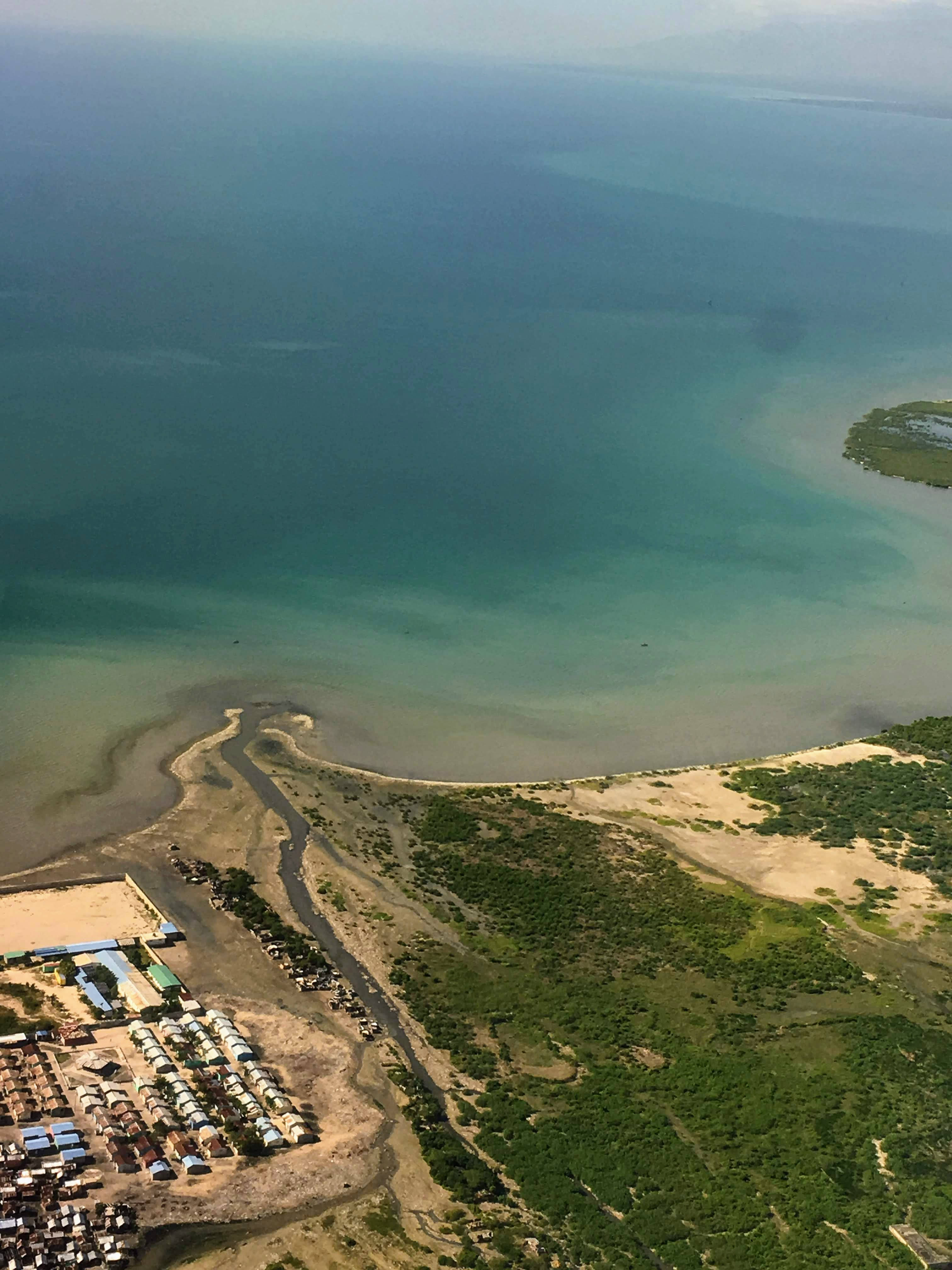 海地空中俯瞰 壁紙 — 俯瞰一條蜿蜒的河流匯入碧綠色的海水，旁邊是一個綠色的沿海定居點，聚集著帳篷和建築物。