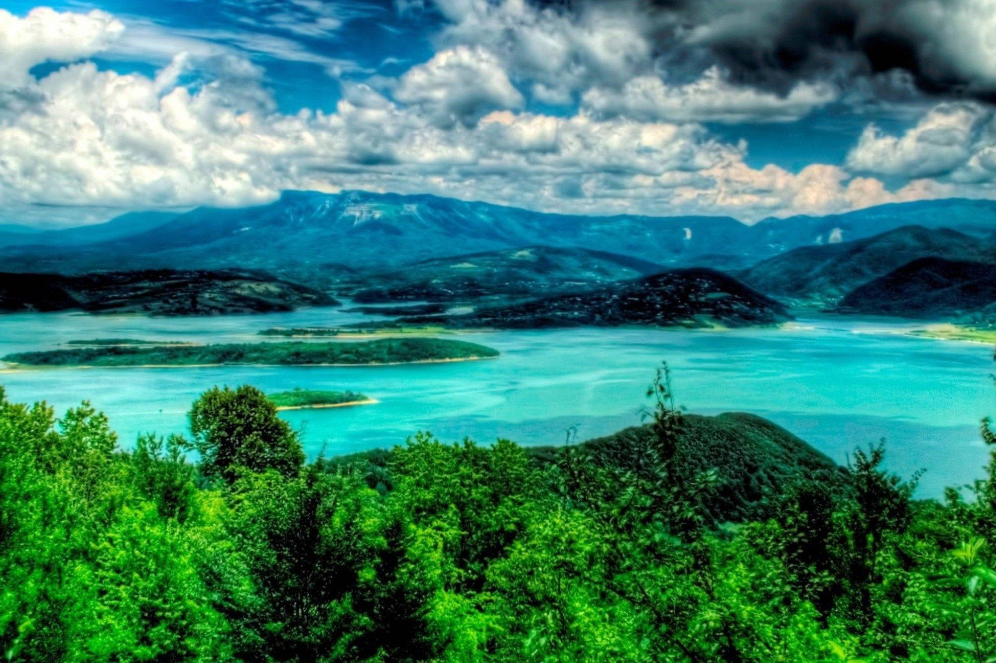 海地絢麗色彩 壁紙 — 在戲劇性的多雲天空下，翠綠的森林和遠山之間鑲嵌著充滿活力的綠松石色湖泊。