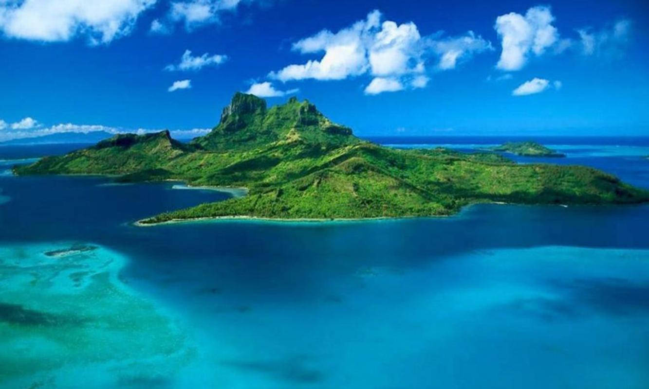 海地色彩繽紛島嶼 壁紙 — 鳥瞰一個鬱鬱蔥蔥的綠色火山島，周圍環繞著充滿活力的碧綠海水，上方是明亮的藍天和蓬鬆的白雲。