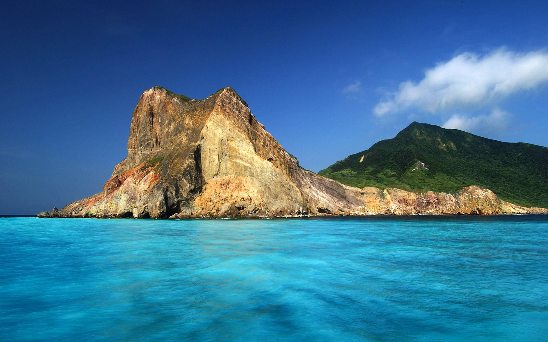 海地陸地與水域 壁紙 — 一座戲劇性的岩石島嶼，帶有層層大地色調，從充滿活力的碧綠海水中升起，天空清澈湛藍，飄著柔軟的白雲。