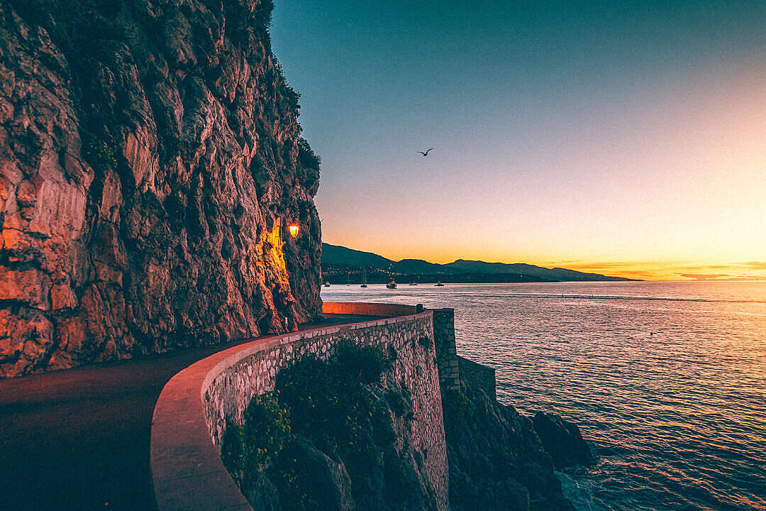 海岸公路日出自然風景 壁紙 — 一條蜿蜒的海岸公路在日落時分緊貼懸崖，溫暖的燈光在清涼的藍色和橙色天空中閃耀。