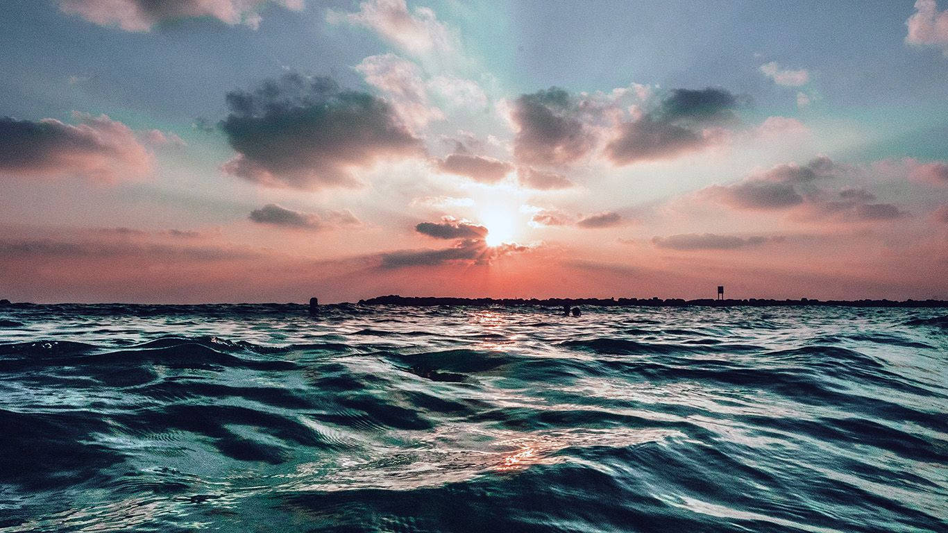 海岸晨 glory 美景 壁紙 — 夕陽穿過雲層，照耀在波光粼粼的海面上，遠處有陸地輪廓。