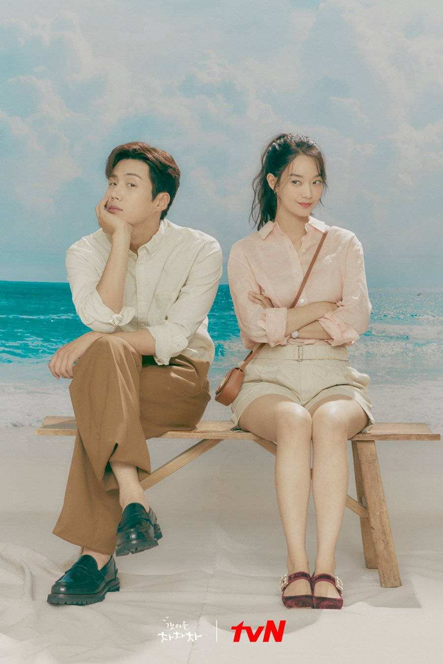《海岸村恰恰恰》海報 壁紙 — 一男一女坐在海邊的木長椅上，穿著粉彩色的夏季服裝，天空湛藍，白雲朵朵。