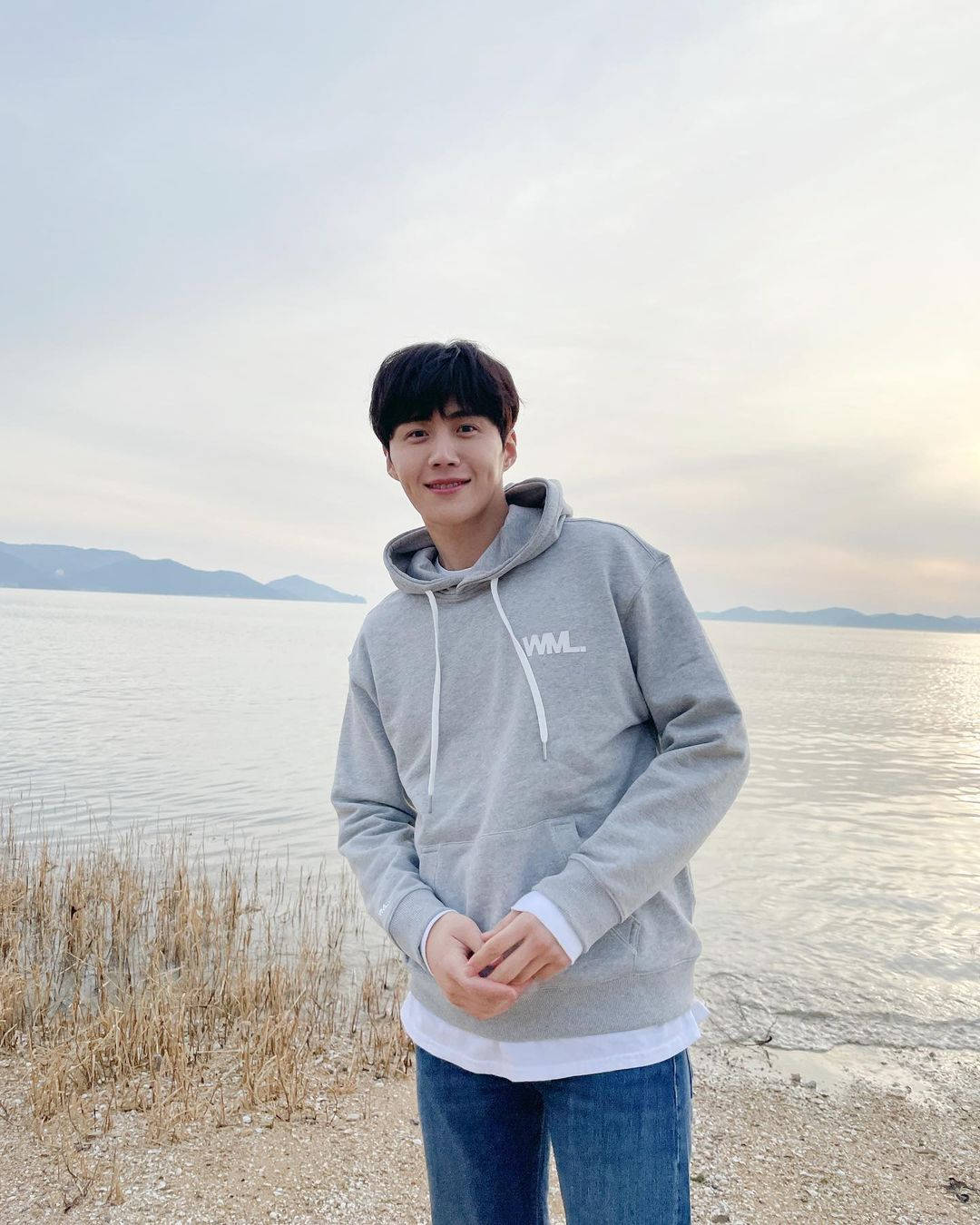 《海岸村恰恰恰》療癒海濱風情 壁紙 — 一位身穿灰色連帽衫的年輕男子在平靜的湖邊微笑，遠處有山巒，天空柔和暮色。