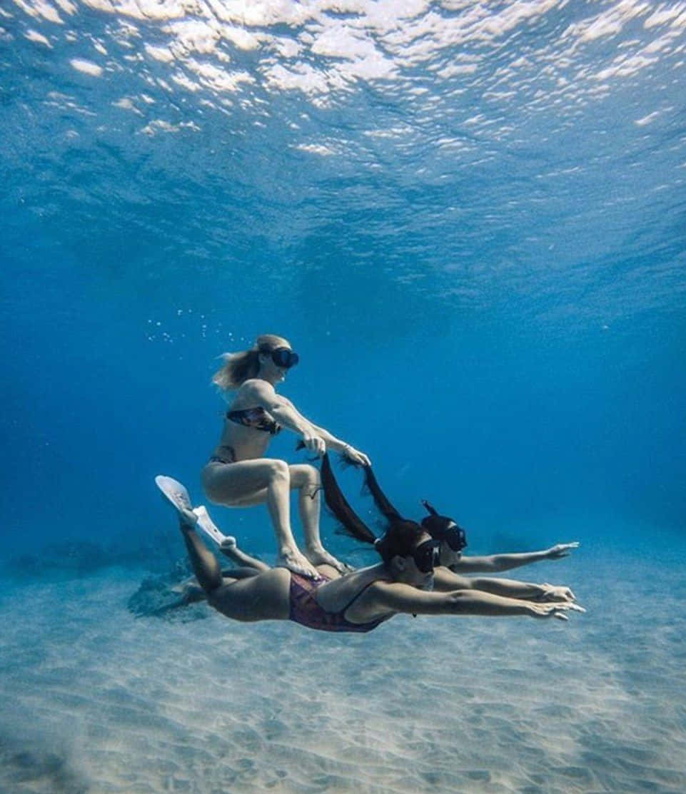 海底嬉戲的女孩們 壁紙 — 兩名穿著浮潛裝備的女性在沙質海床上優雅地滑行，其中一人騎在另一人身上。