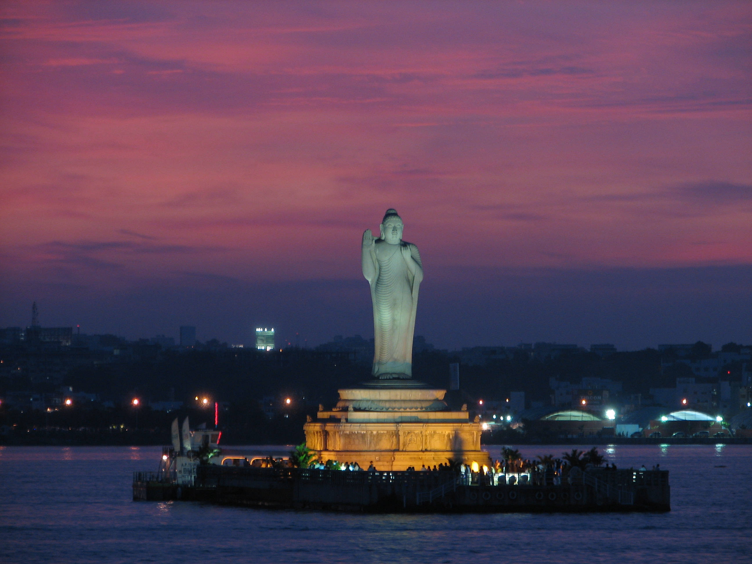 海得拉巴薩加湖夜景 壁紙 — 一座高大的佛像在充滿活力的粉紅色和紫色暮色天空下，在水濱平台上被照亮。