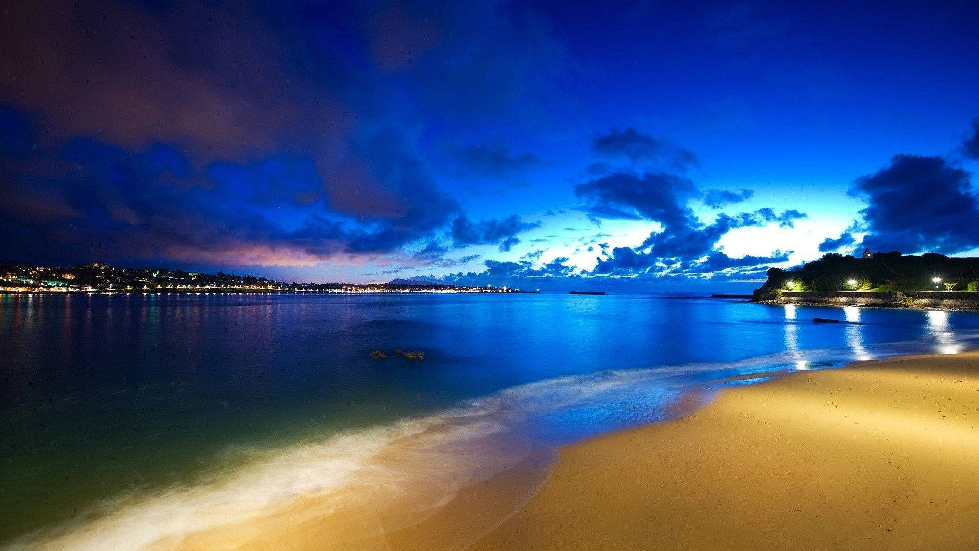 海景 Windows Vista 壁紙 — 一個寧靜的黃昏海灘，有金色沙灘、平靜的藍色海水，以及在戲劇性的多雲天空下反射的遠處城市燈光。