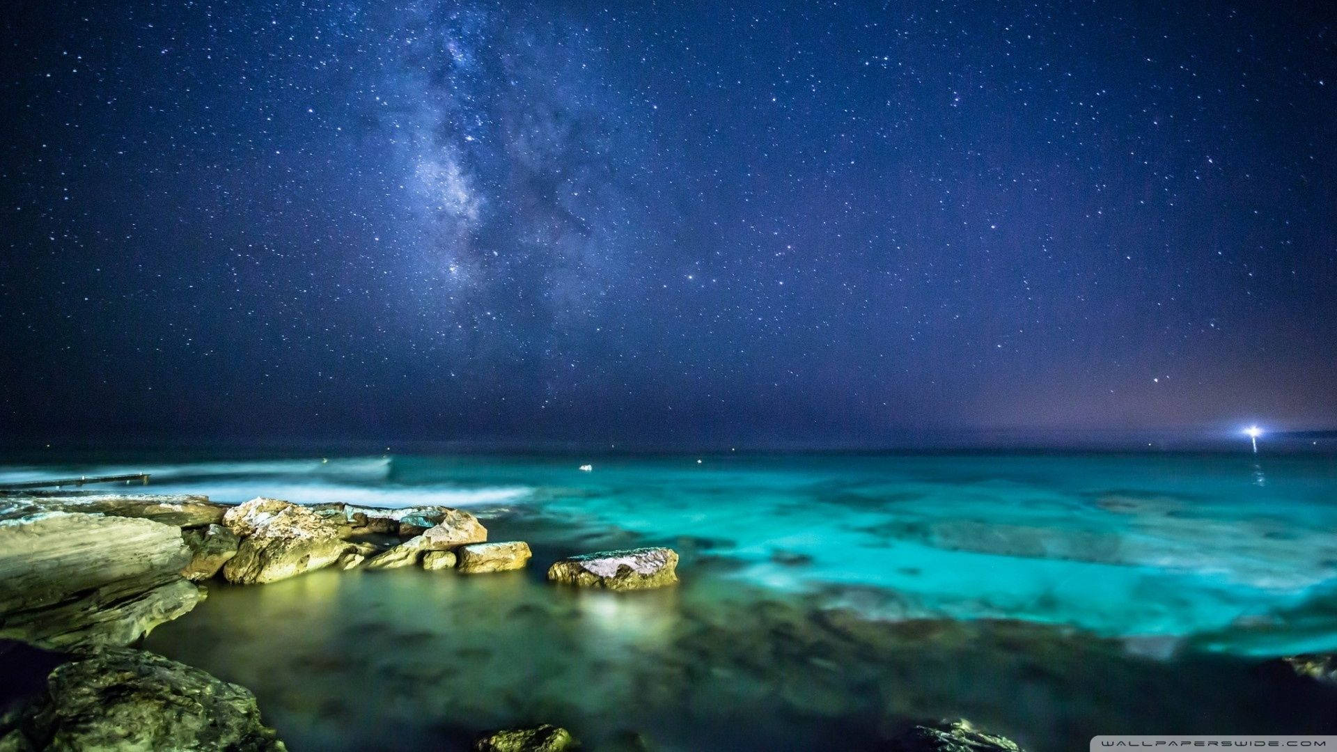繁星點點的夜空，銀河在閃爍的藍綠色海洋和岩石海岸上方。