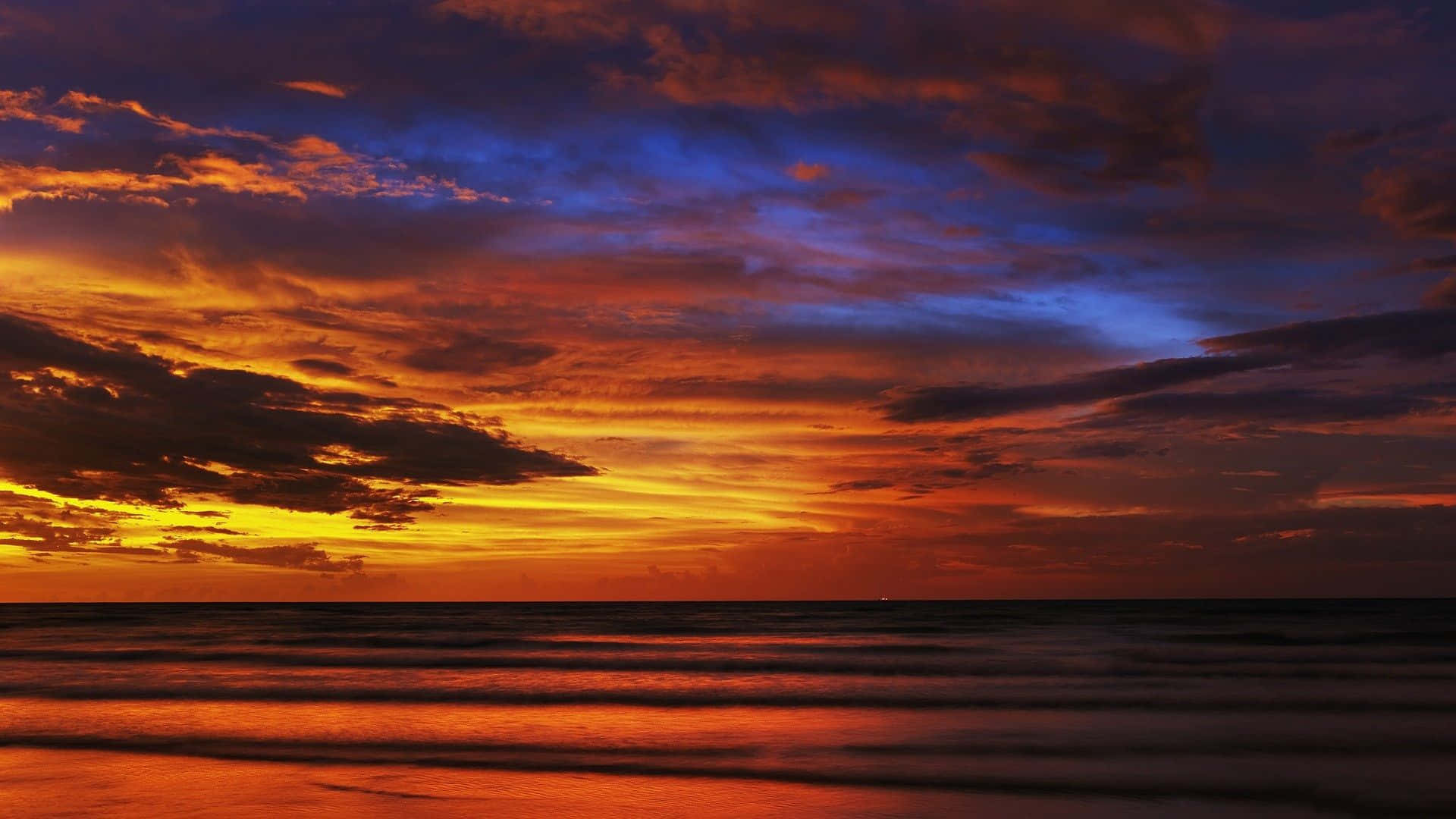Sunset View 桌布 壁紙 — 海洋上空戲劇性的日落，火橙色和深藍色的雲彩映照在濕沙上。