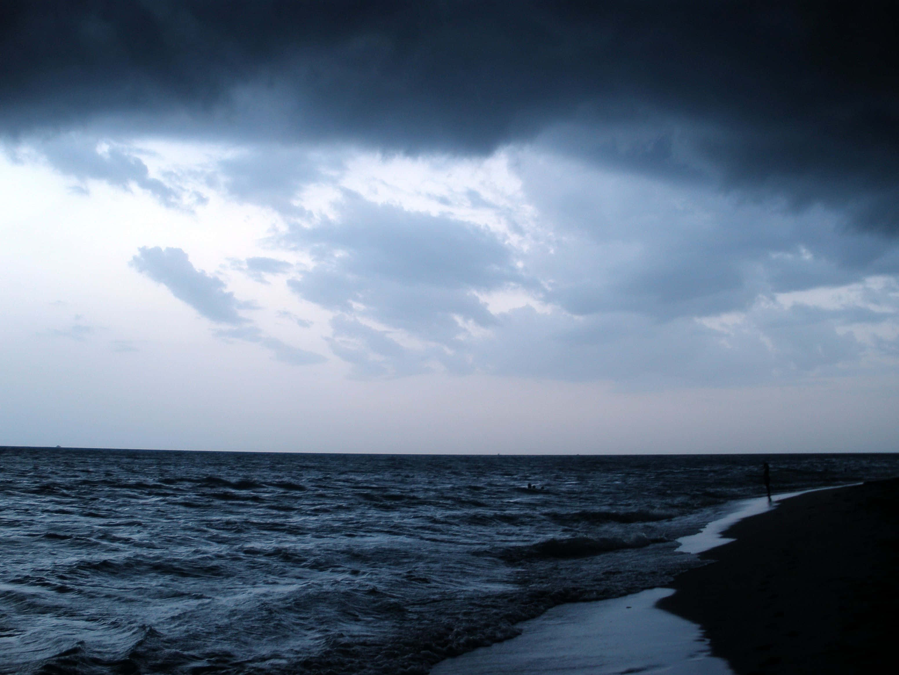 海洋上空的壯觀暴風雲 壁紙 — 一個孤獨的身影沿著黑暗的海灘行走，暴風雲在暮色天空下籠罩著洶湧的海浪。
