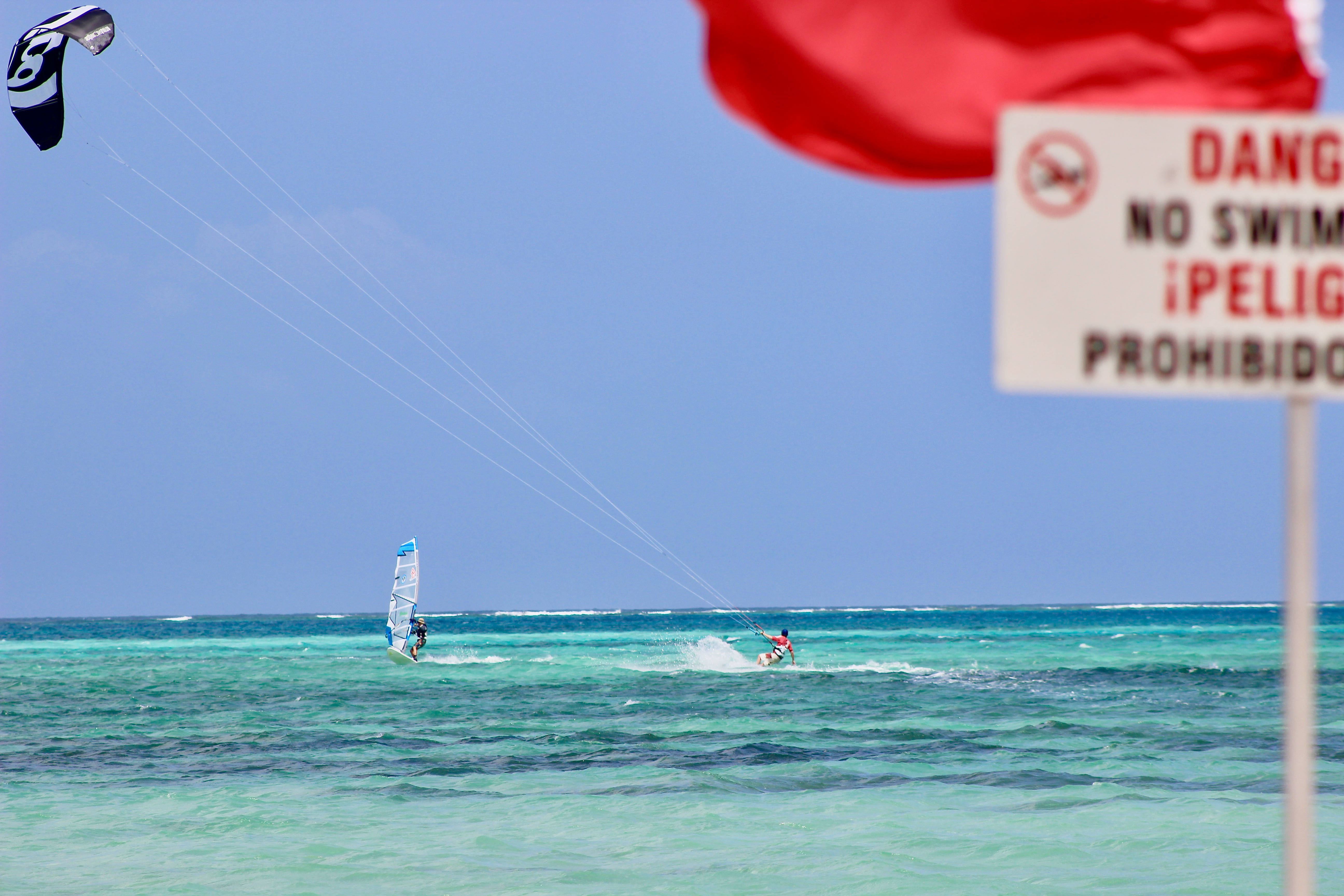 海洋中的風帆衝浪區域 壁紙 — 風帆衝浪者和風箏衝浪者在晴朗藍天下，於碧綠海水上滑行，附近有紅旗和警告標誌。