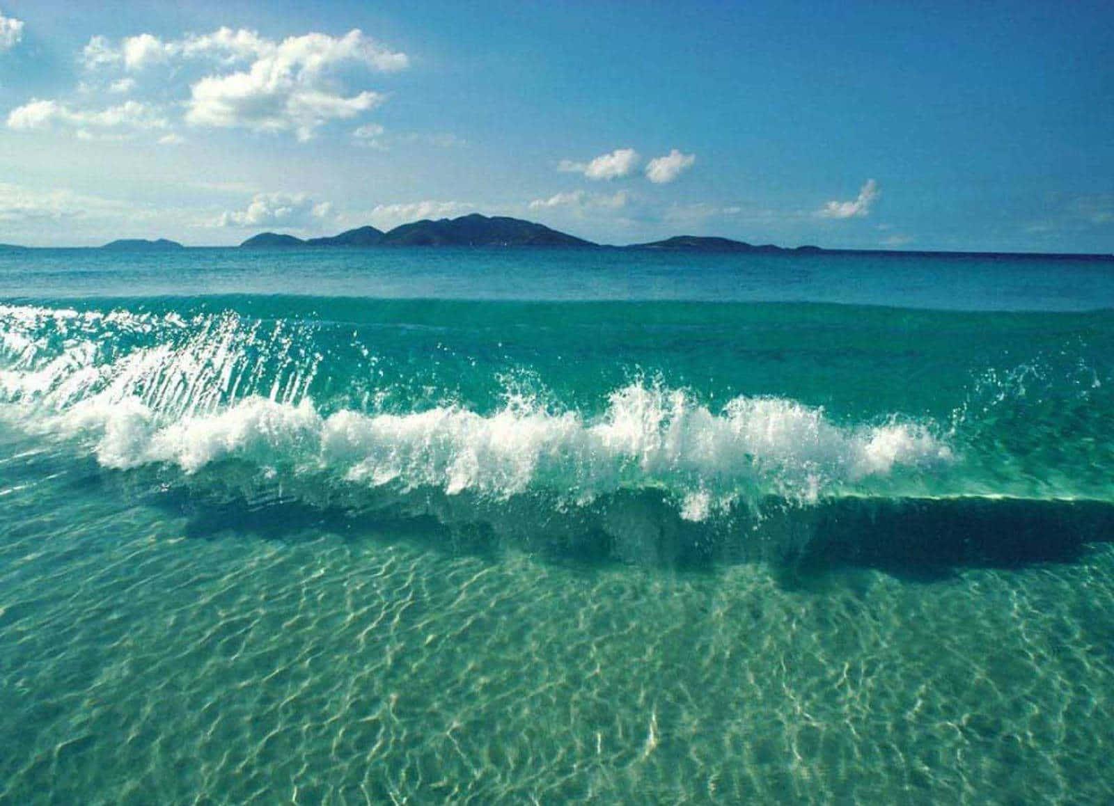 海洋之美 壁紙 — 充滿活力的藍綠色波浪帶著白色泡沫拍打著清澈的藍天和遠處的島嶼山脈。