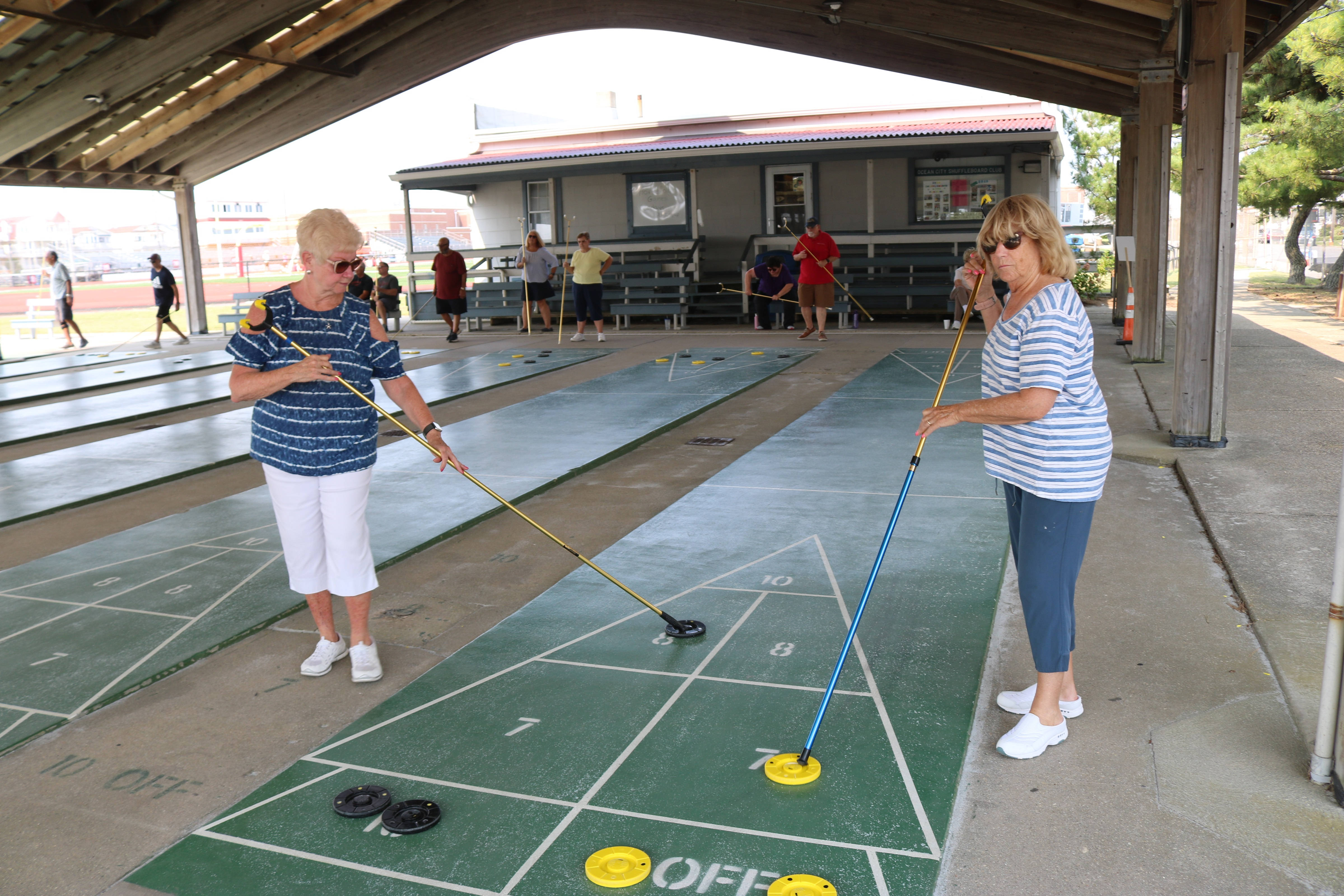 海洋城 Shuffleboard 遊戲比賽 壁紙 — 兩位身穿條紋襯衫的女性在陽光明媚的日子裡，在木製涼亭下玩沙狐球。