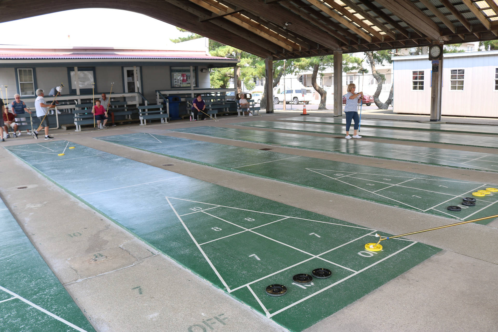 海洋城體育複合體 Shuffleboard 球場 壁紙 — 玩家在木製涼亭下進行沙狐球比賽，場地為綠色，碟片散落各處。