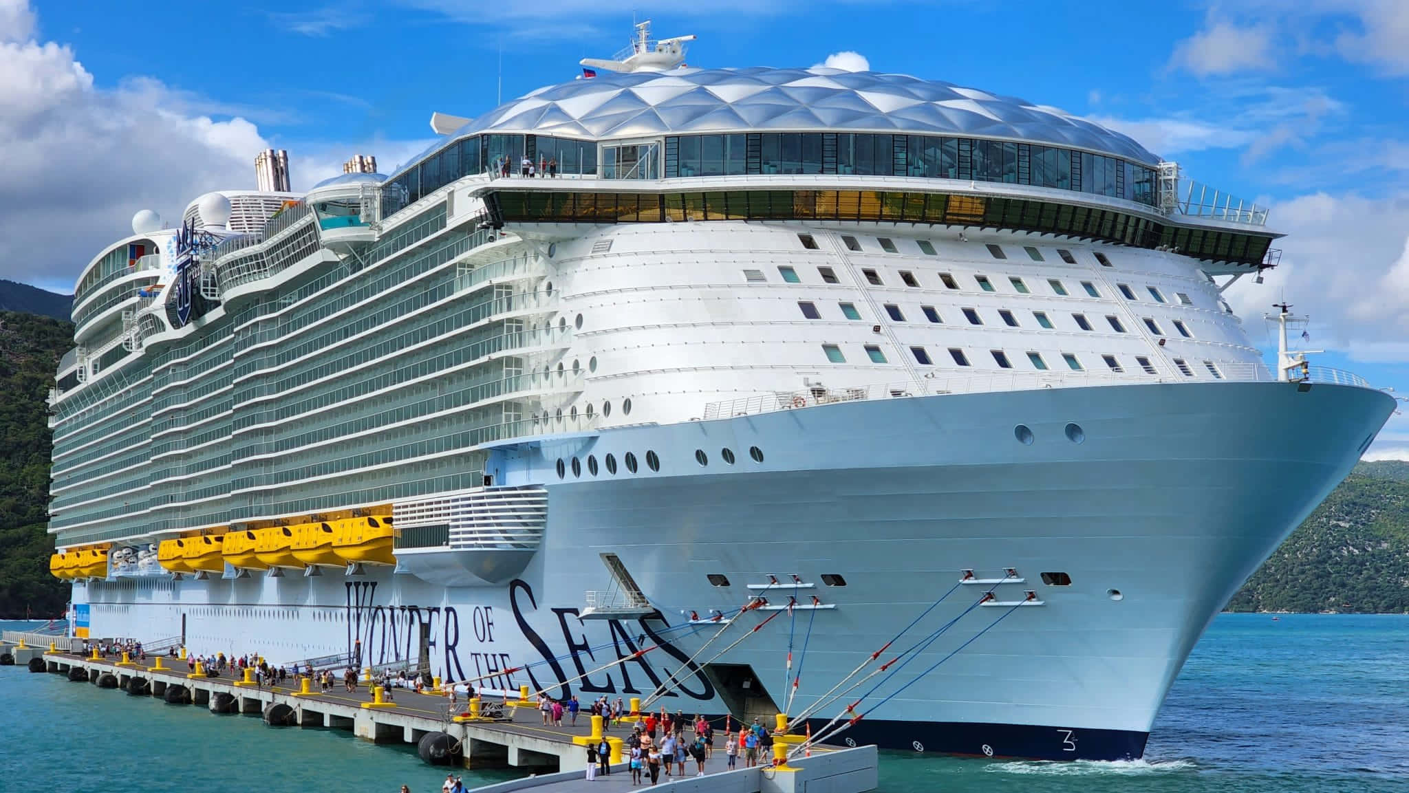 Cruise Ship 桌布 壁紙 — 海洋奇蹟號郵輪停靠在一個熱帶港口，周圍是碧綠的海水和鬱鬱蔥蔥的綠色山丘，頭頂是明亮的藍天。
