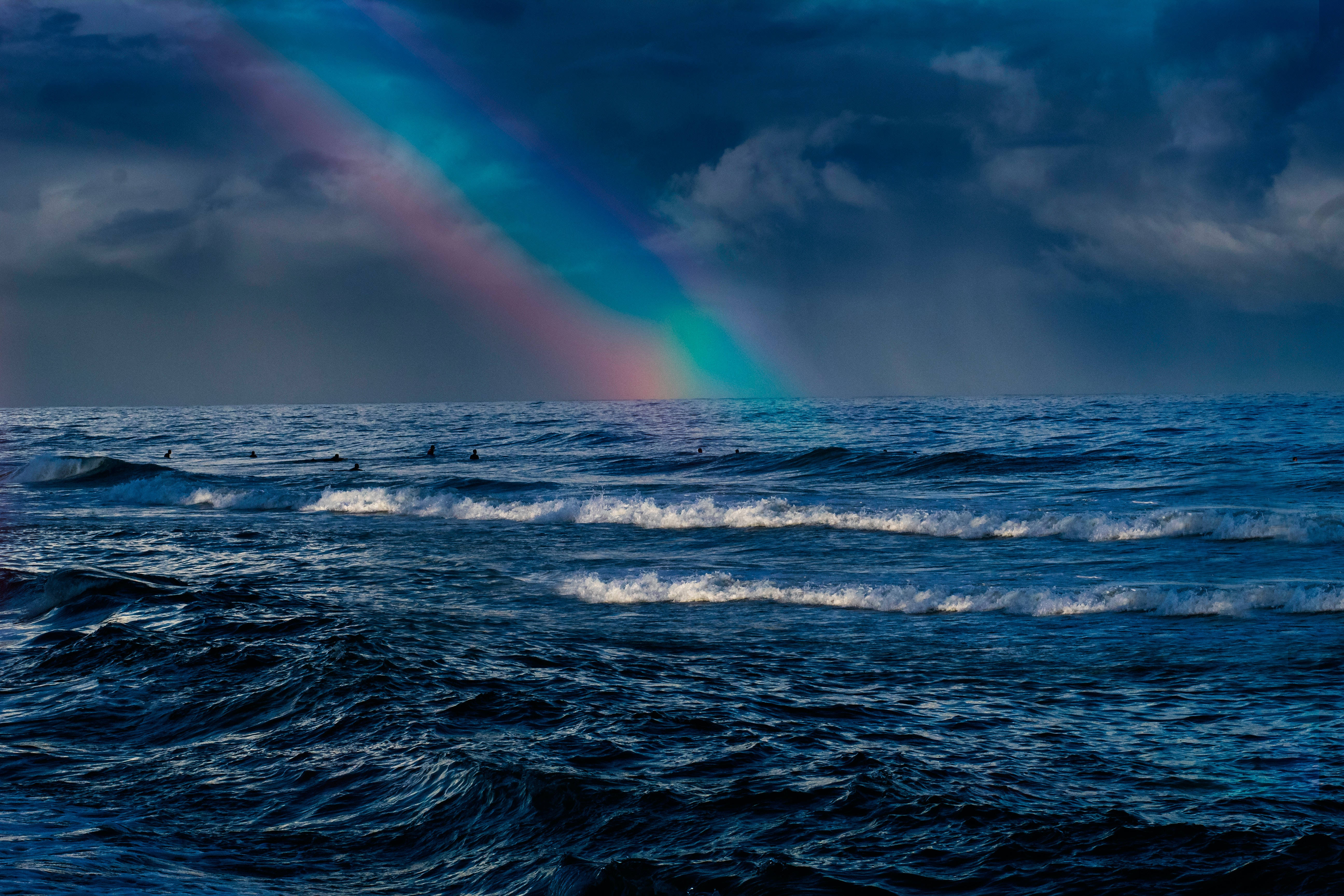 海洋彩虹 4K 壁紙 — 一道戲劇性的彩虹橫跨暴風雨中的海洋，衝浪者在烏雲下乘風破浪。
