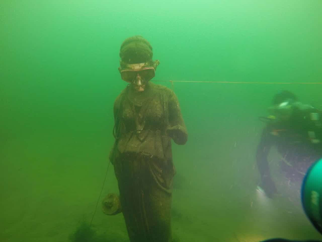 海洋的黑暗深處 壁紙 — 一座長滿青苔的雕像浸在渾濁的綠色水中，附近有潛水員在探索。